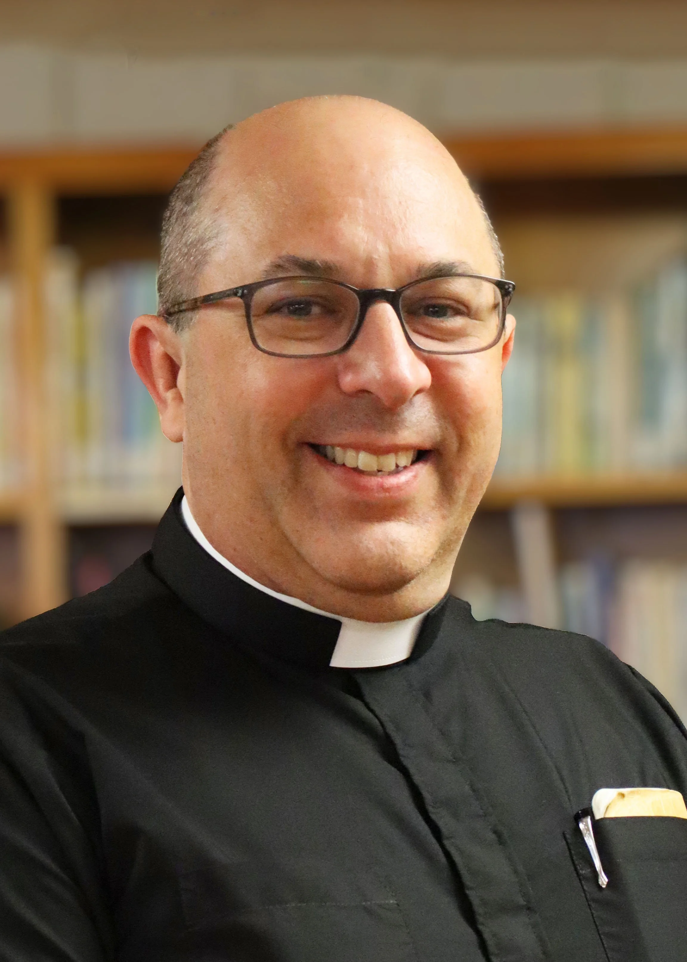 Close-up of a smiling man wearing glasses and a black clerical shirt with a white clerical collar, possibly a priest, in front of a background of bookshelves.