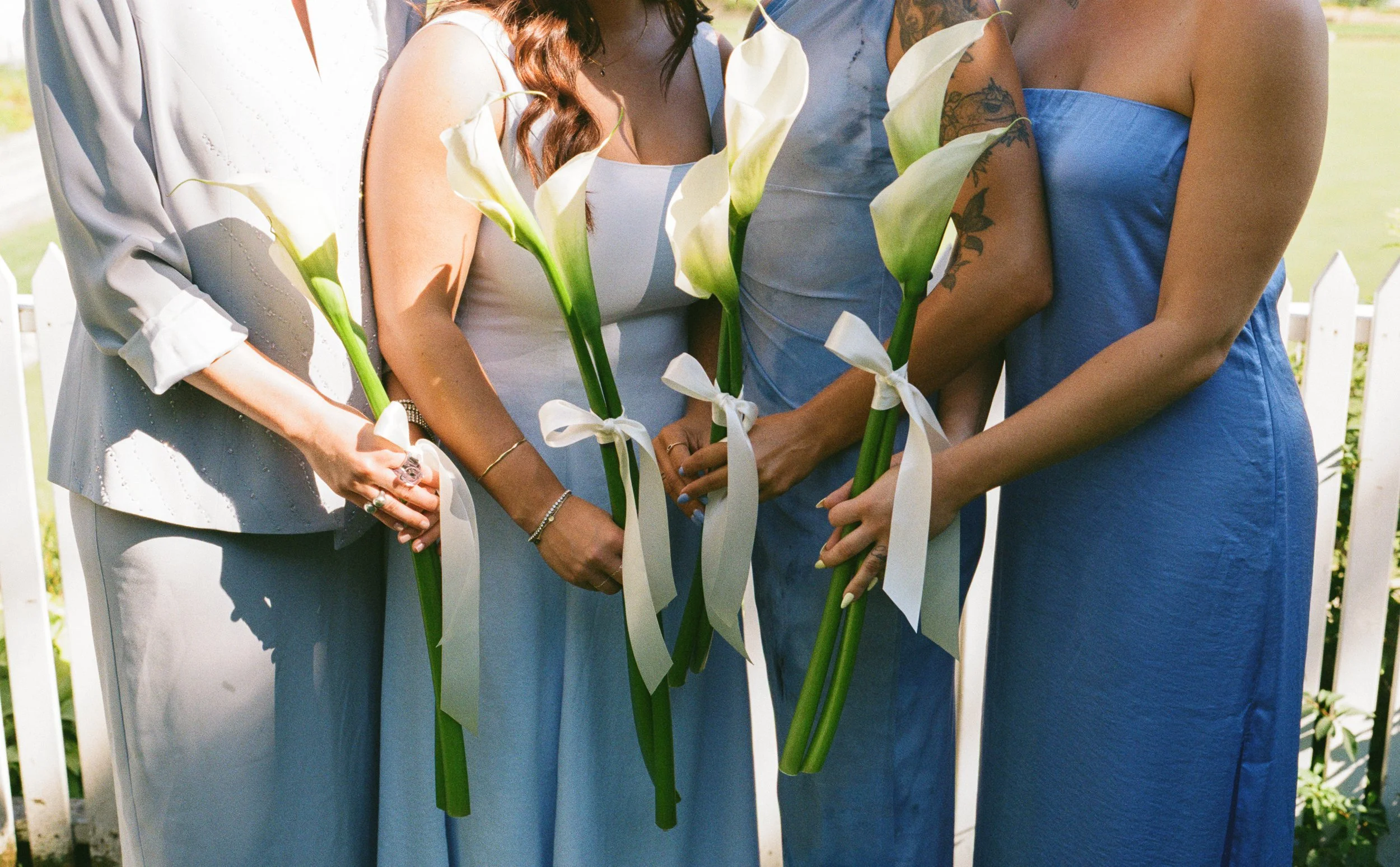 Close-up of four women holding white calla lilies with tied ribbons, wearing blue and white dresses, outdoors behind a white picket fence.