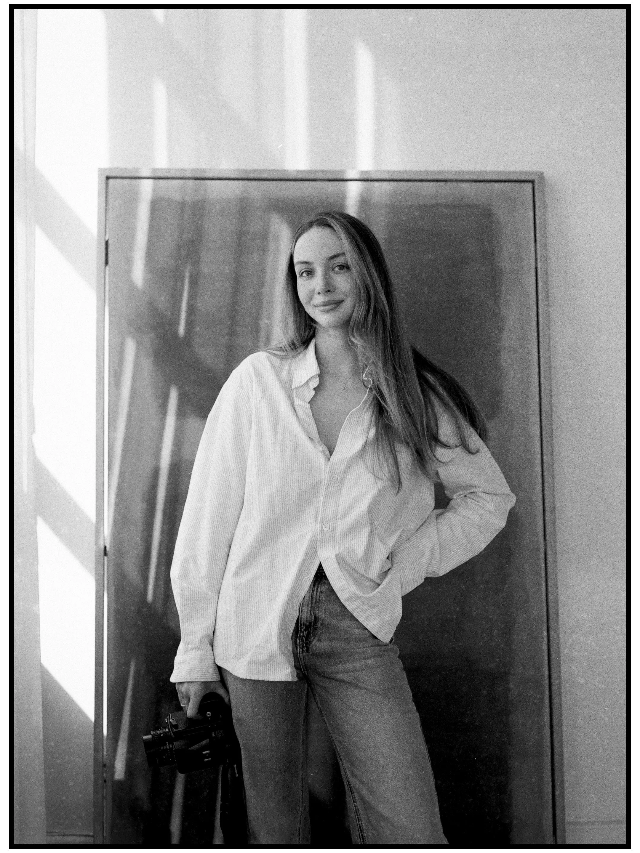Black and white photo of a young woman with long hair, wearing a button-up shirt and jeans, standing in front of a chalkboard, holding a camera in her left hand.