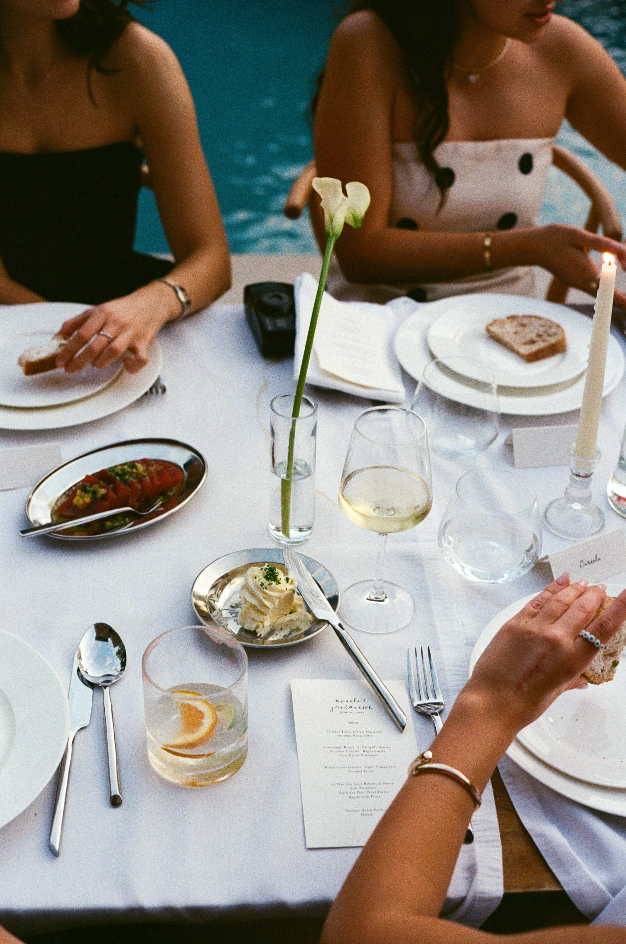 A table set for a meal with four women, with plates of bread, a dish of lobster, a glass of white wine, a glass of water with lemon, and a single flower in a vase, near a pool.