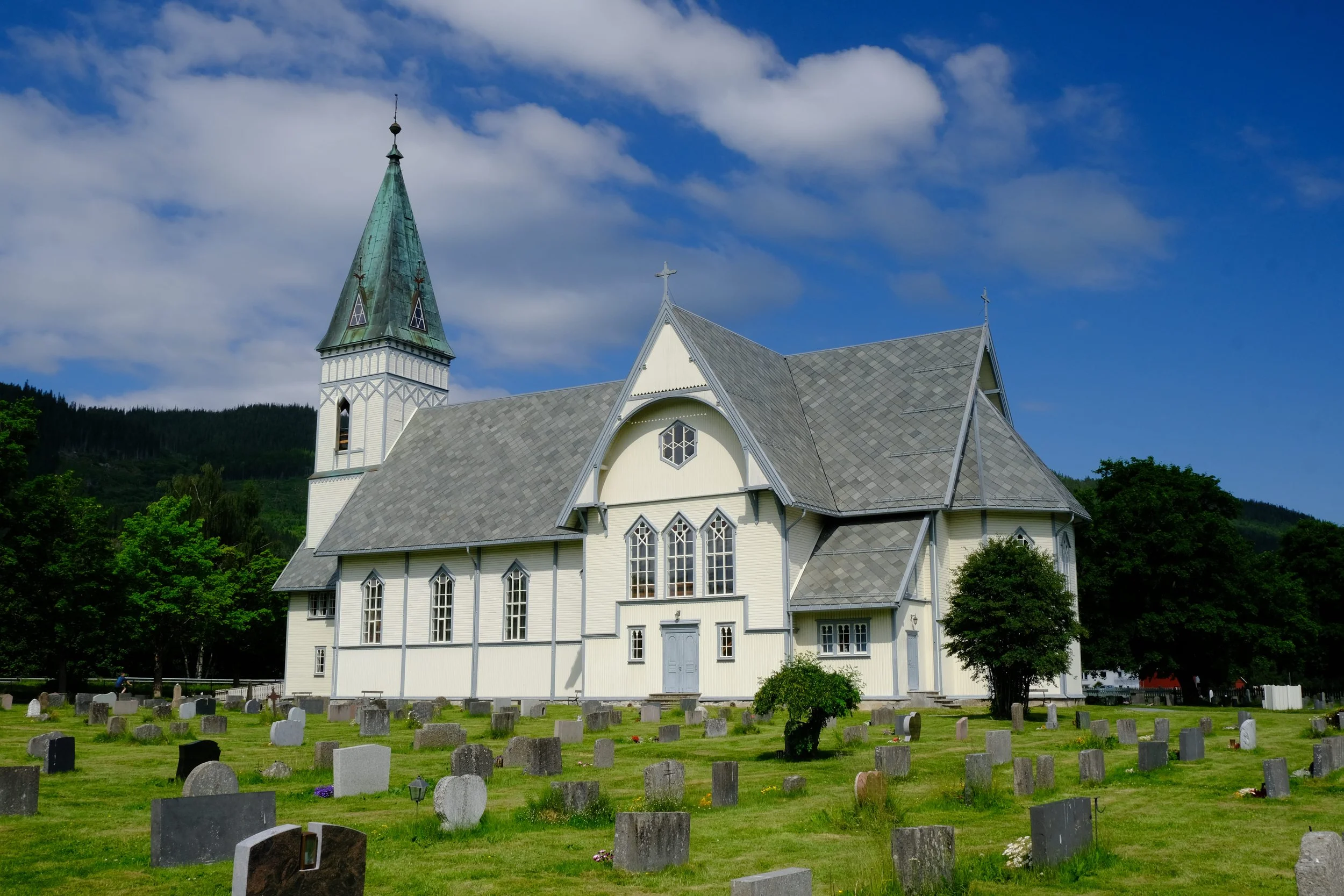 Eine weiße Kirche mit Spitzdach und Glockenturm, umgeben von einem Friedhof mit Grabsteinen, vor einem blauen Himmel mit Wolken, im Grünen mit Bäumen und Hügeln im Hintergrund.