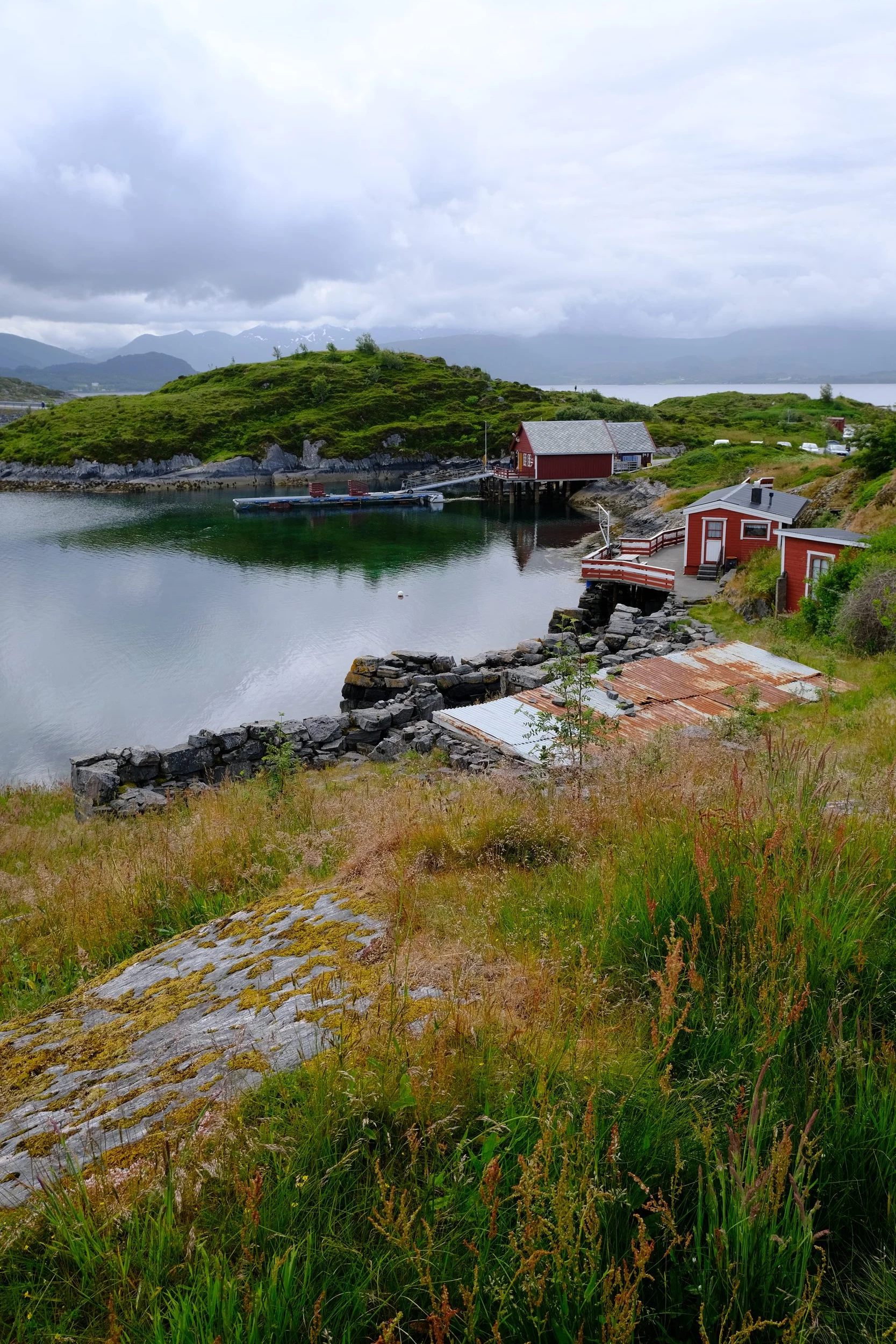 Blick auf ein ruhiges Hafenörtchen mit roten Holzhäusern, grüner Insel im Hintergrund und bewölktem Himmel