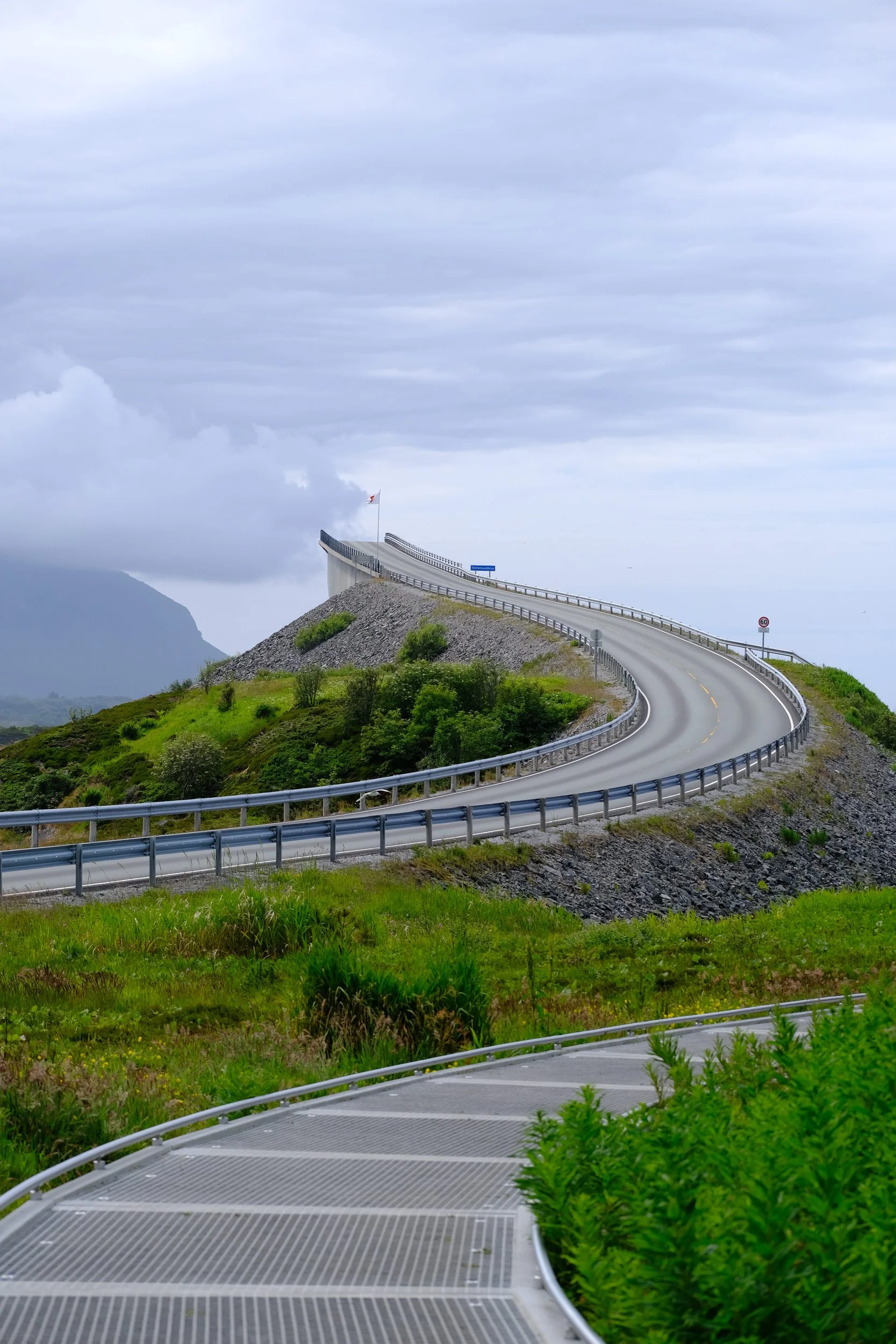 Kurvige Straße in bergiger Landschaft mit grünen Pflanzen und Wolken am Himmel.