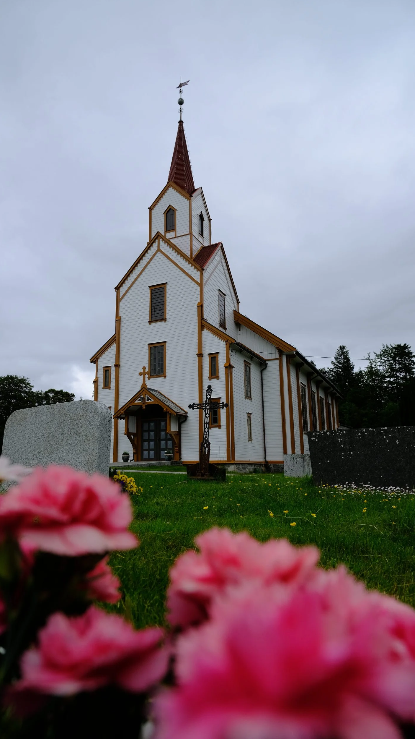 Eine weiße Kirche mit roten Dachspitzen, ein Kreuz vor der Kirche, im Vordergrund pinke Blumen, bewölkter Himmel.
