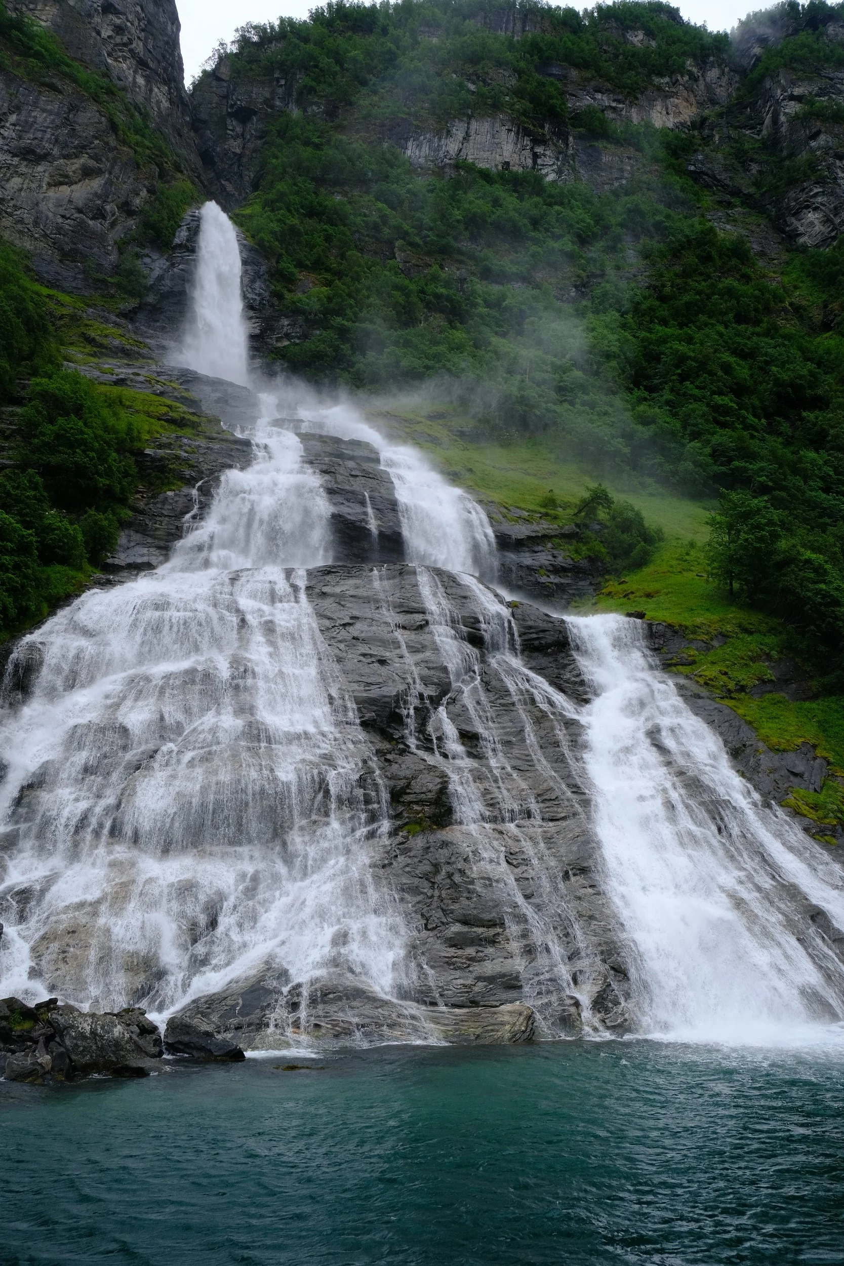 Mehrstufiger Wasserfall inmitten grüner Berge, Wasser stürzt auf einen Fjord