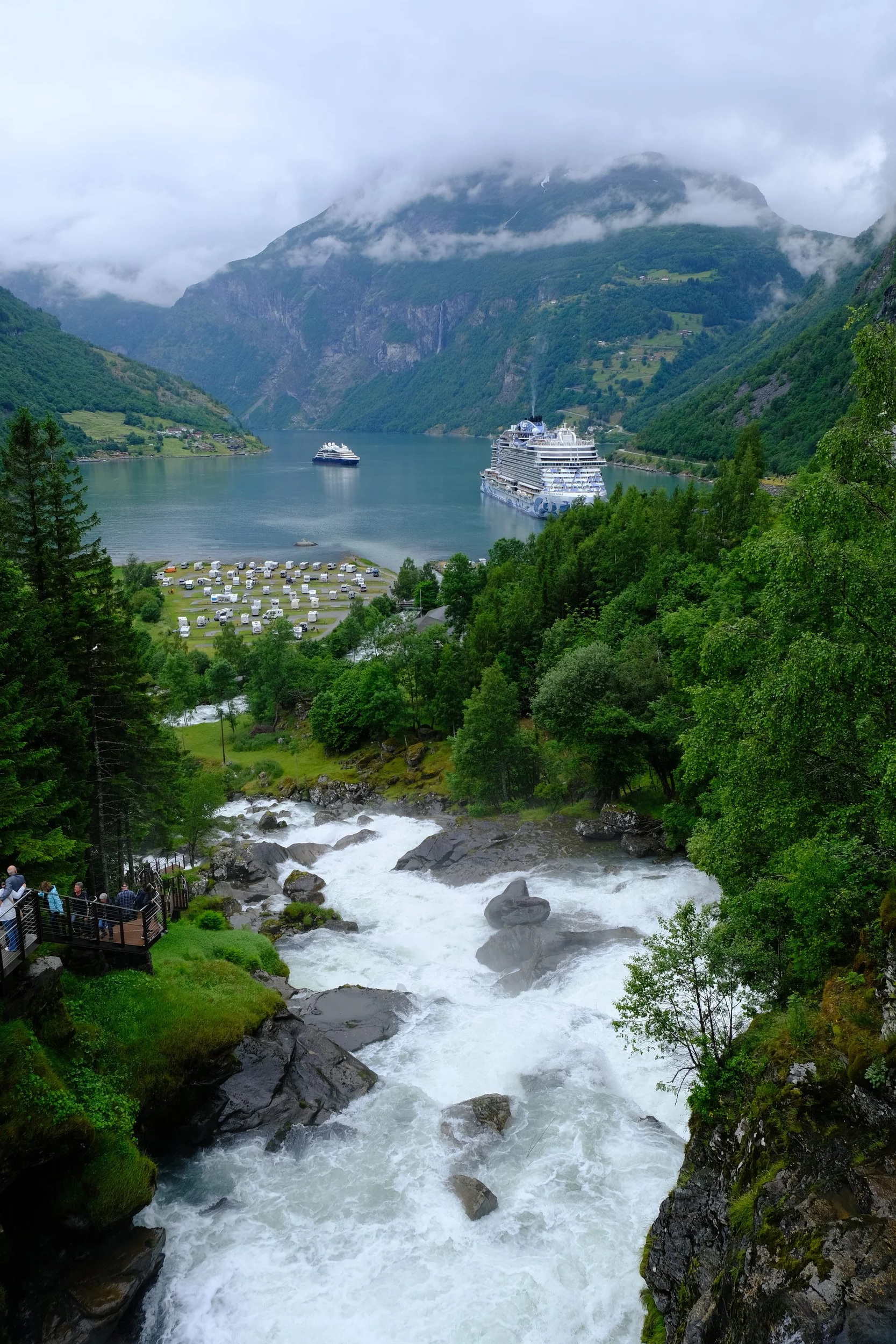 Blick auf einen Fjord mit schneebedecktem Gebirge im Hintergrund. Im Wasser sind zwei Kreuzfahrtschiffe. Im Vordergrund fließt ein Wasserfall durch eine grüne, bewaldete Landschaft. Es gibt einen Aussichtspunkt mit mehreren Menschen.
