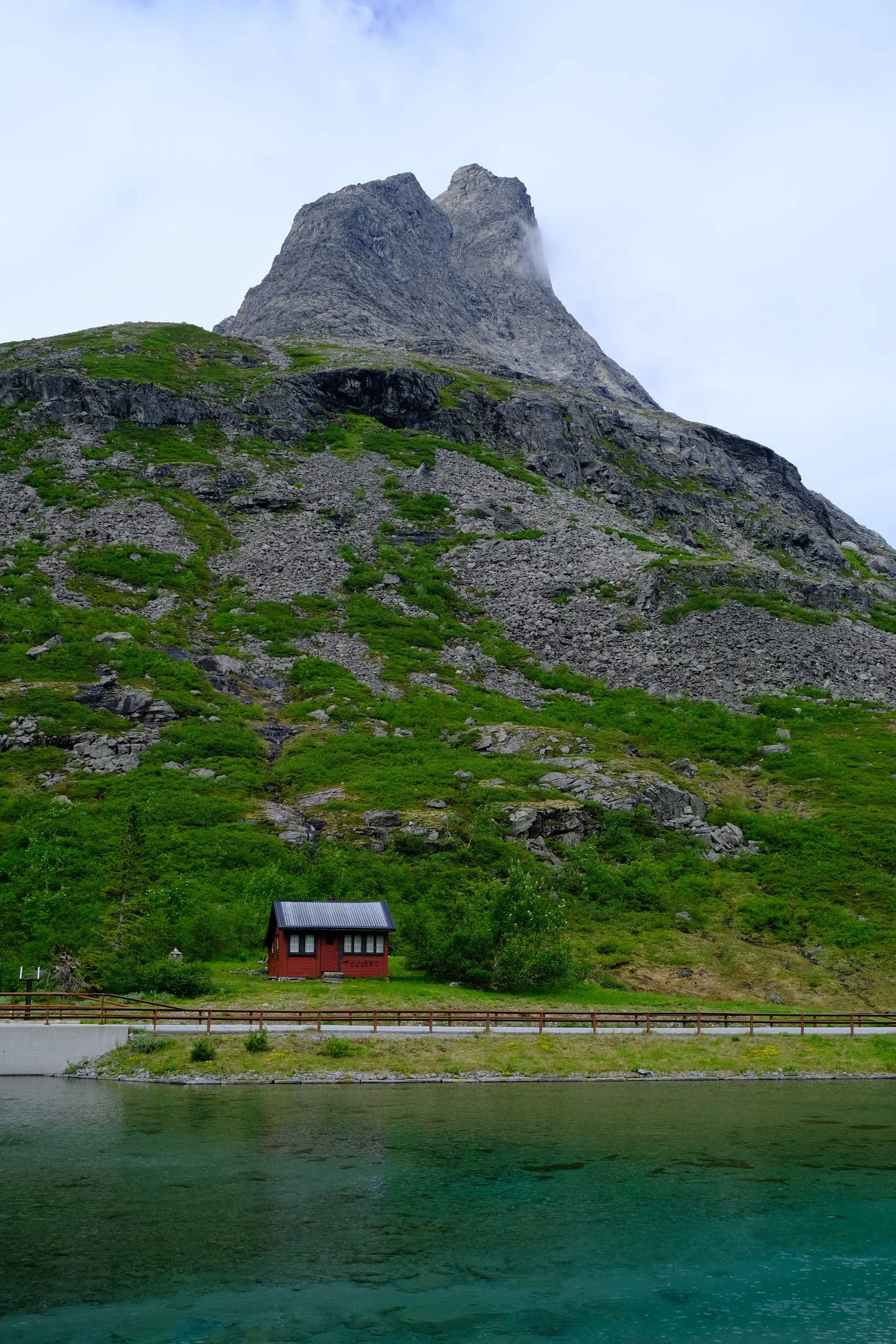 Berge mit grünem Vegetation, kleine rote Hütte, Wasser im Vordergrund, bewölkter Himmel.