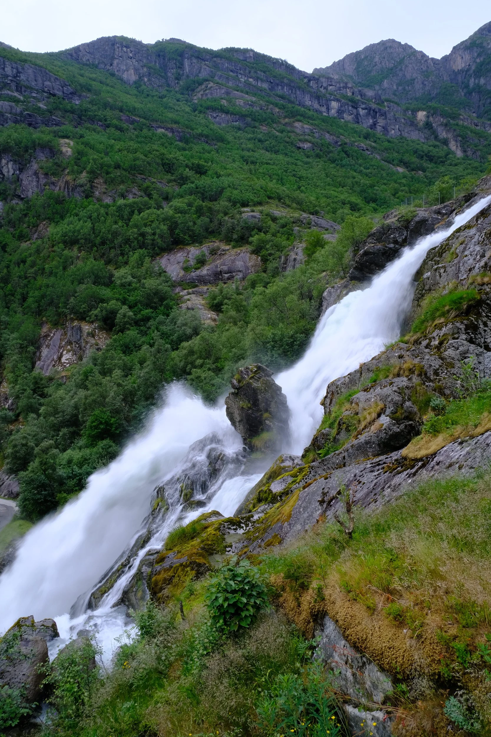 Ein Wasserfall stürzt den felsigen Hang hinab, umgeben von grünen Bäumen und Bergen.