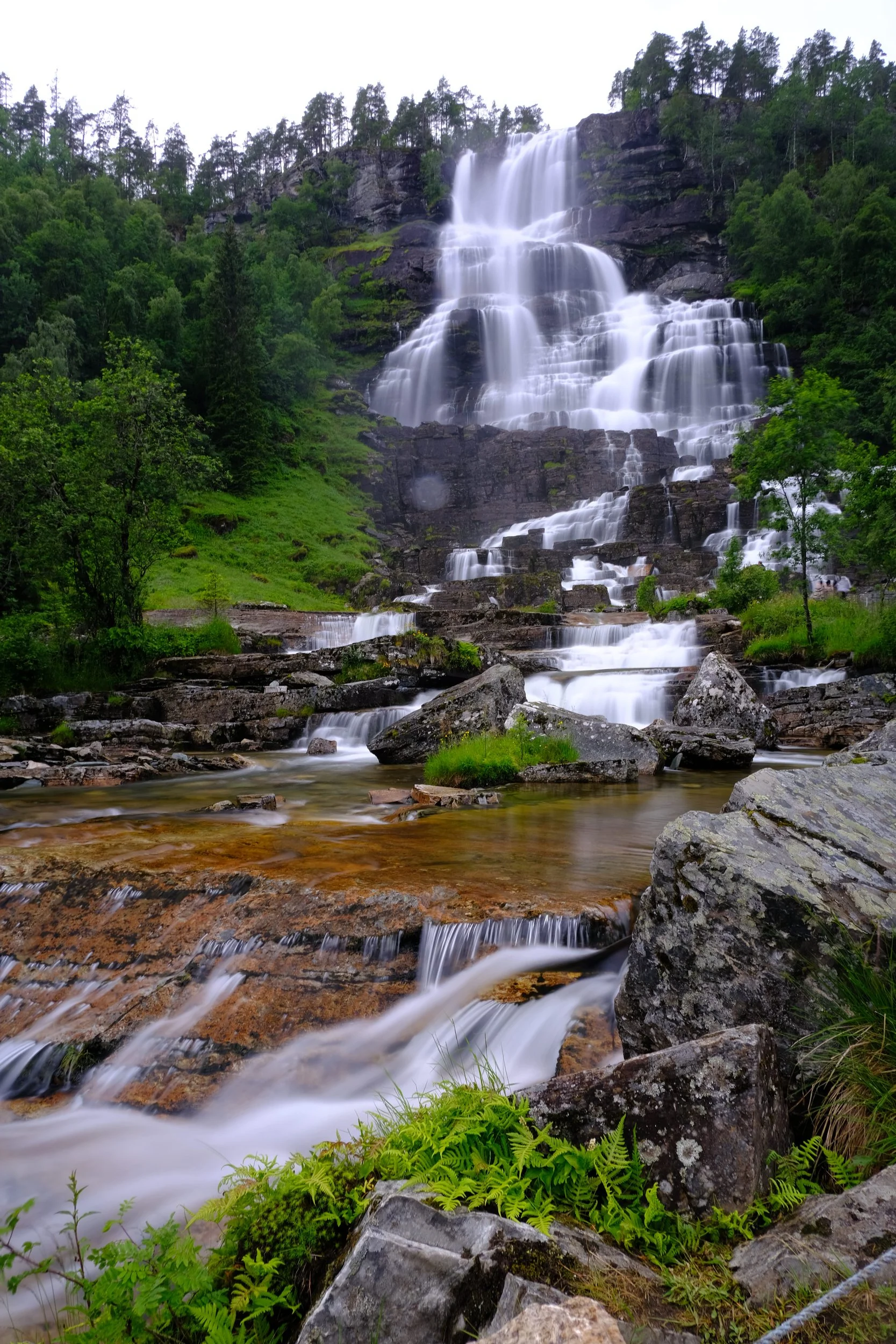 Mehrstufiger Wasserfall inmitten einer grünen, bewaldeten Landschaft mit Felsen.