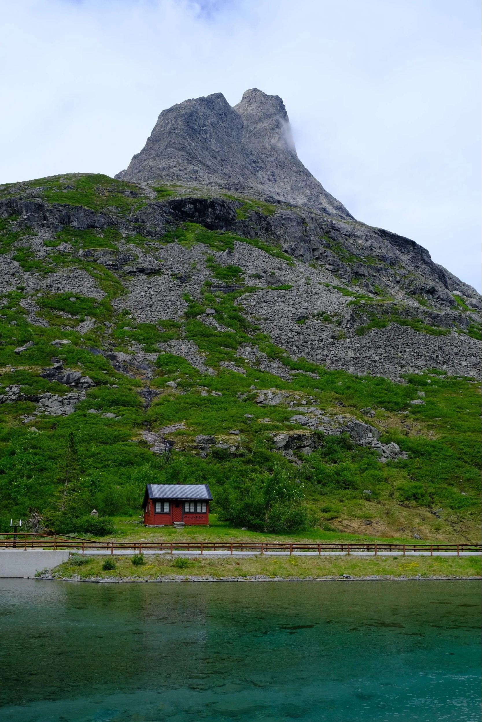 Ein Haus am Ufer eines Sees mit einem großen, felsigen Berg im Hintergrund. Der Himmel ist bewölkt.