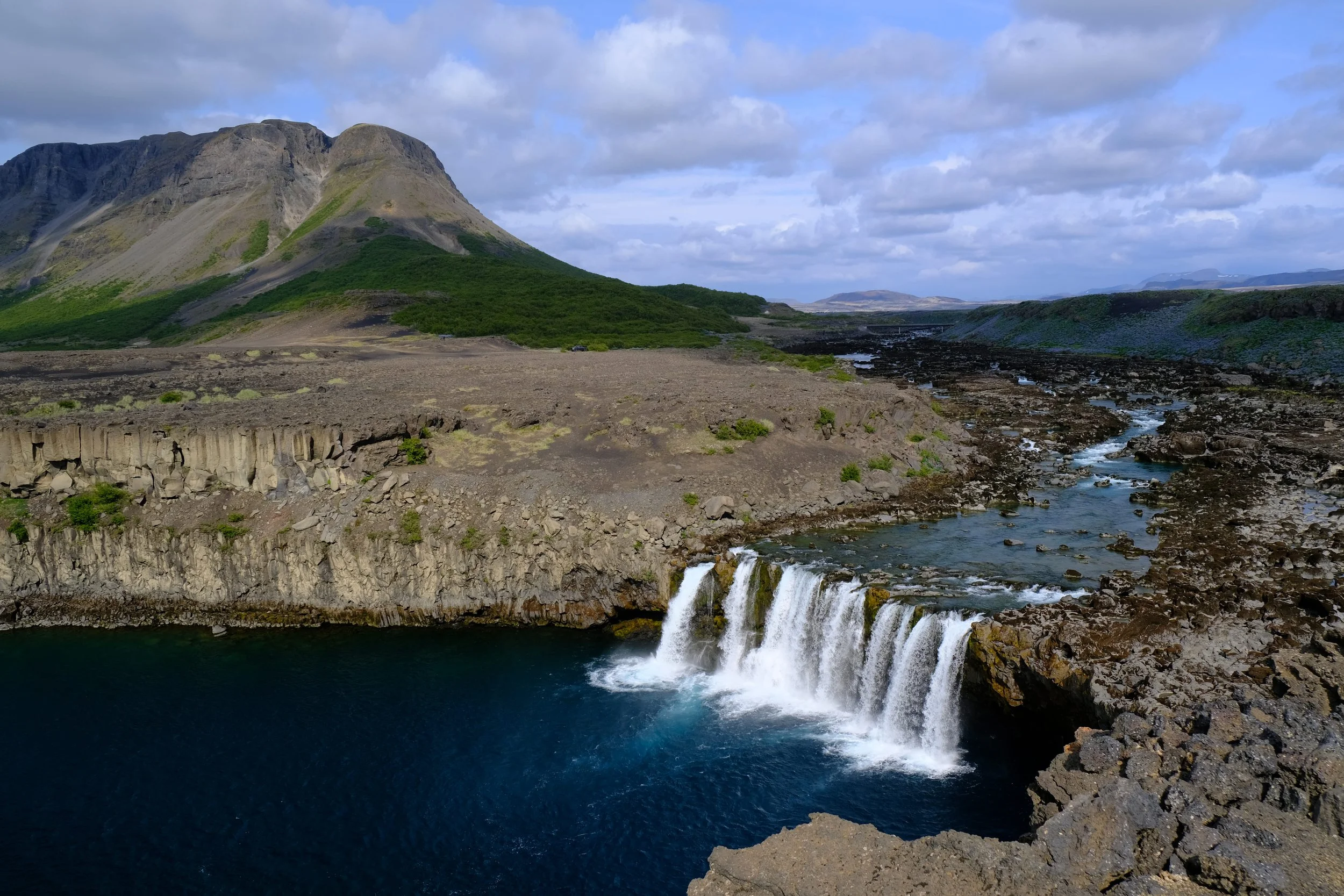 Ein Wasserfall in einer wilden Landschaft mit Bergen im Hintergrund, Wasser fließt in einen dunklen See, Wolken am Himmel.