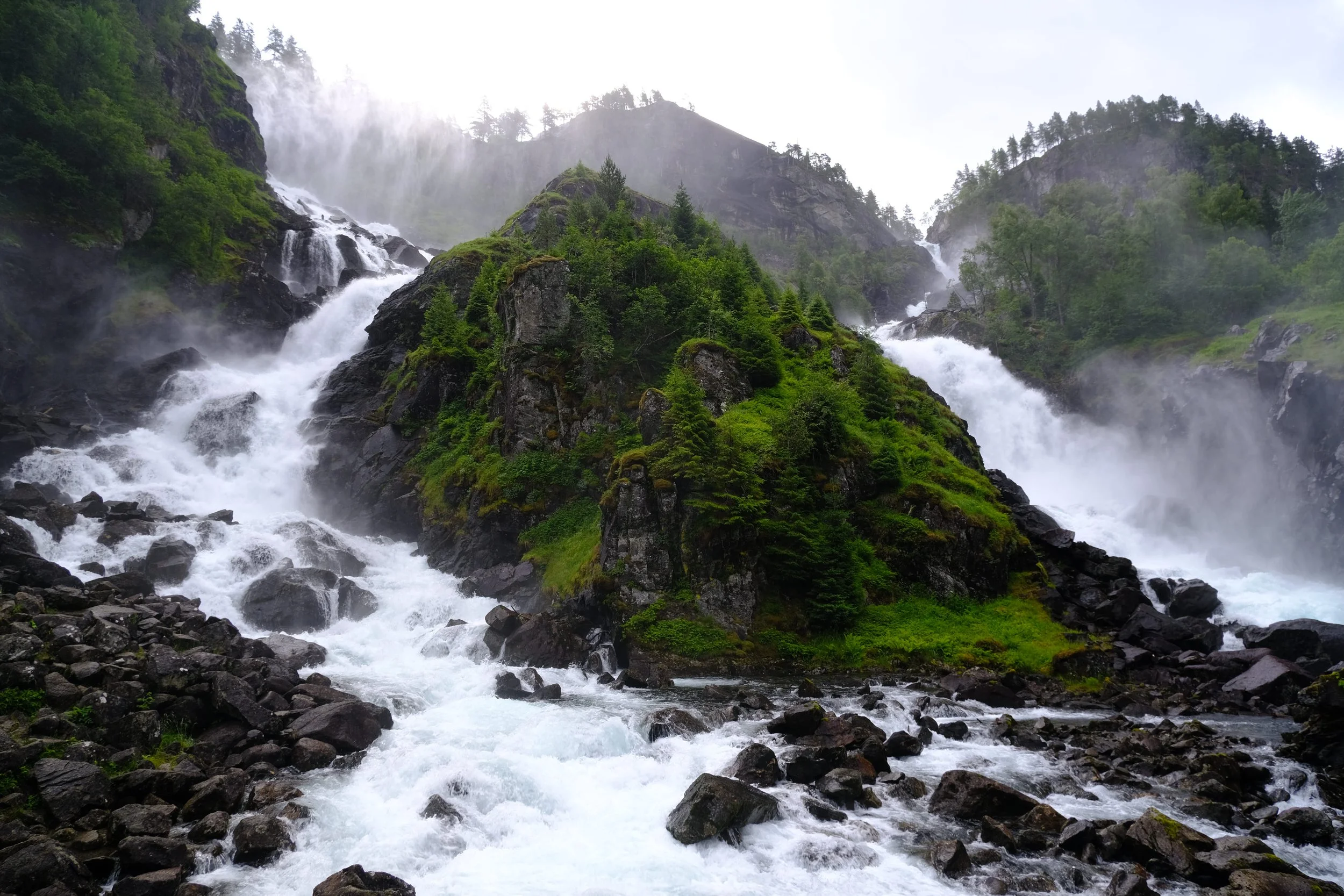 Ein Wasserfall inmitten einer grünen, bewaldeten Berglandschaft mit steinigem Fluss unten.