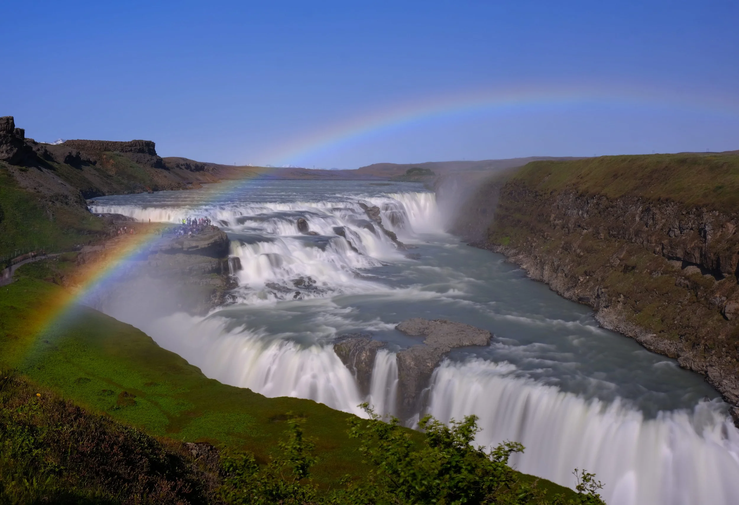 Mehrere Stufen des Wasserfalles Gullfoss in Island mit Regenbogen über dem Wasser und in der Luft, grüne Hänge, blauer Himmel.