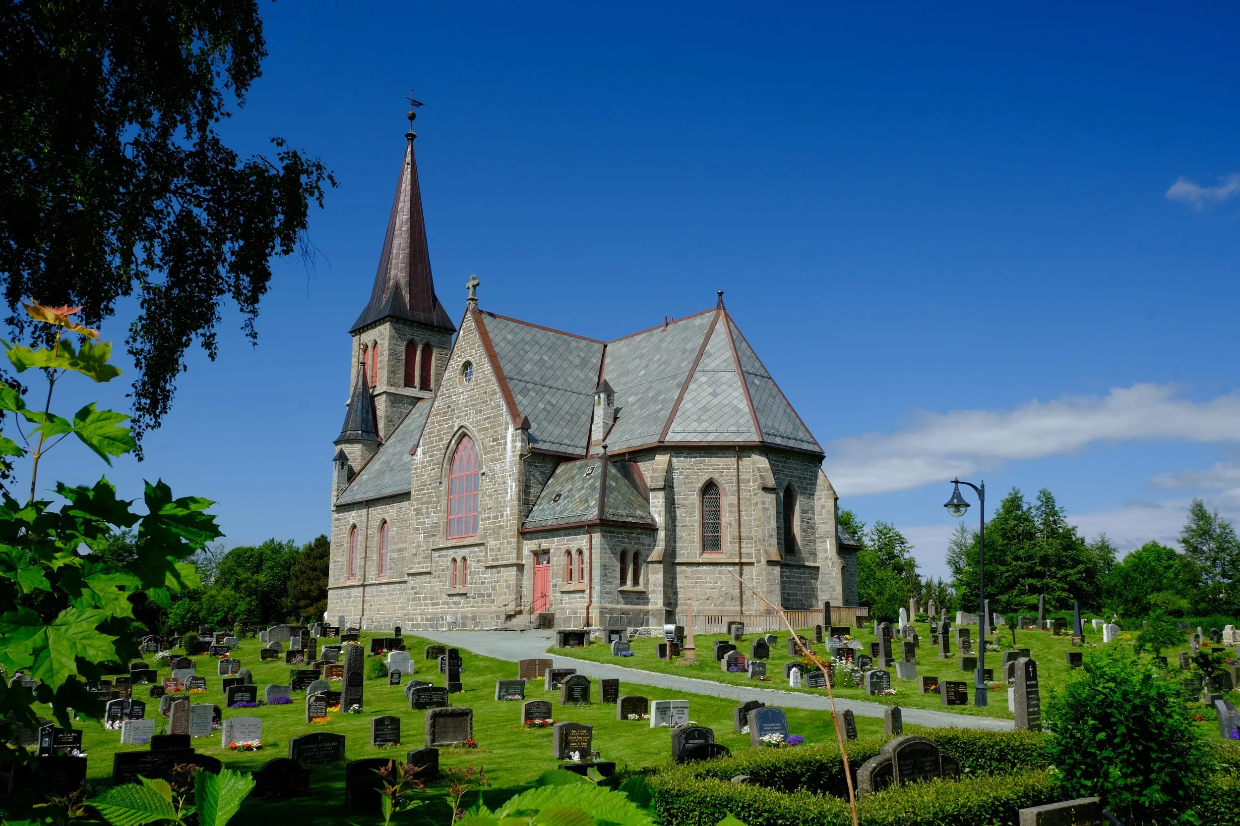 Eine steinerne Kirche mit hohen Fenstern und einem spitzen Turm, umgeben von einem Friedhof mit Grabsteinen, bei sonnigem Himmel.