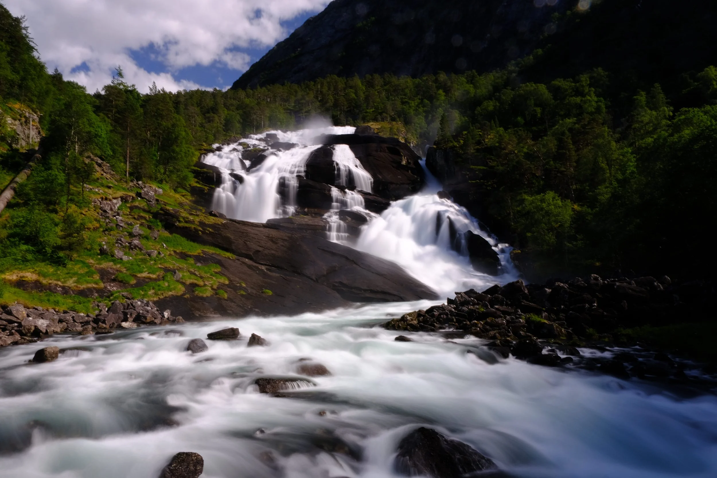 Ein Wasserfall inmitten eines grünen Waldes, umgeben von Bergen und fliessendem Wasser im Vordergrund.