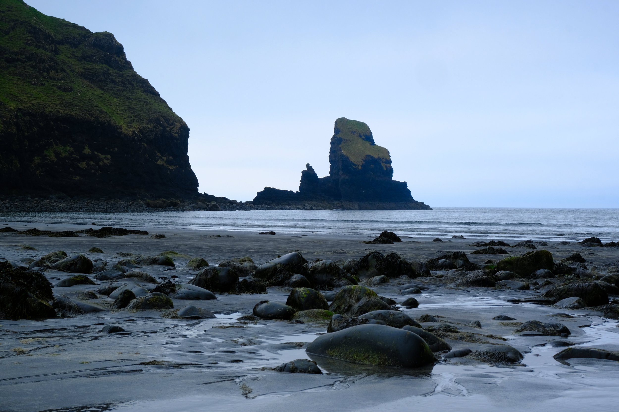 Ein Strand mit dunklem Sand und vielen glatten Steinen, eine Klippe im Hintergrund und eine Felsformation im Wasser vor dem Horizont.
