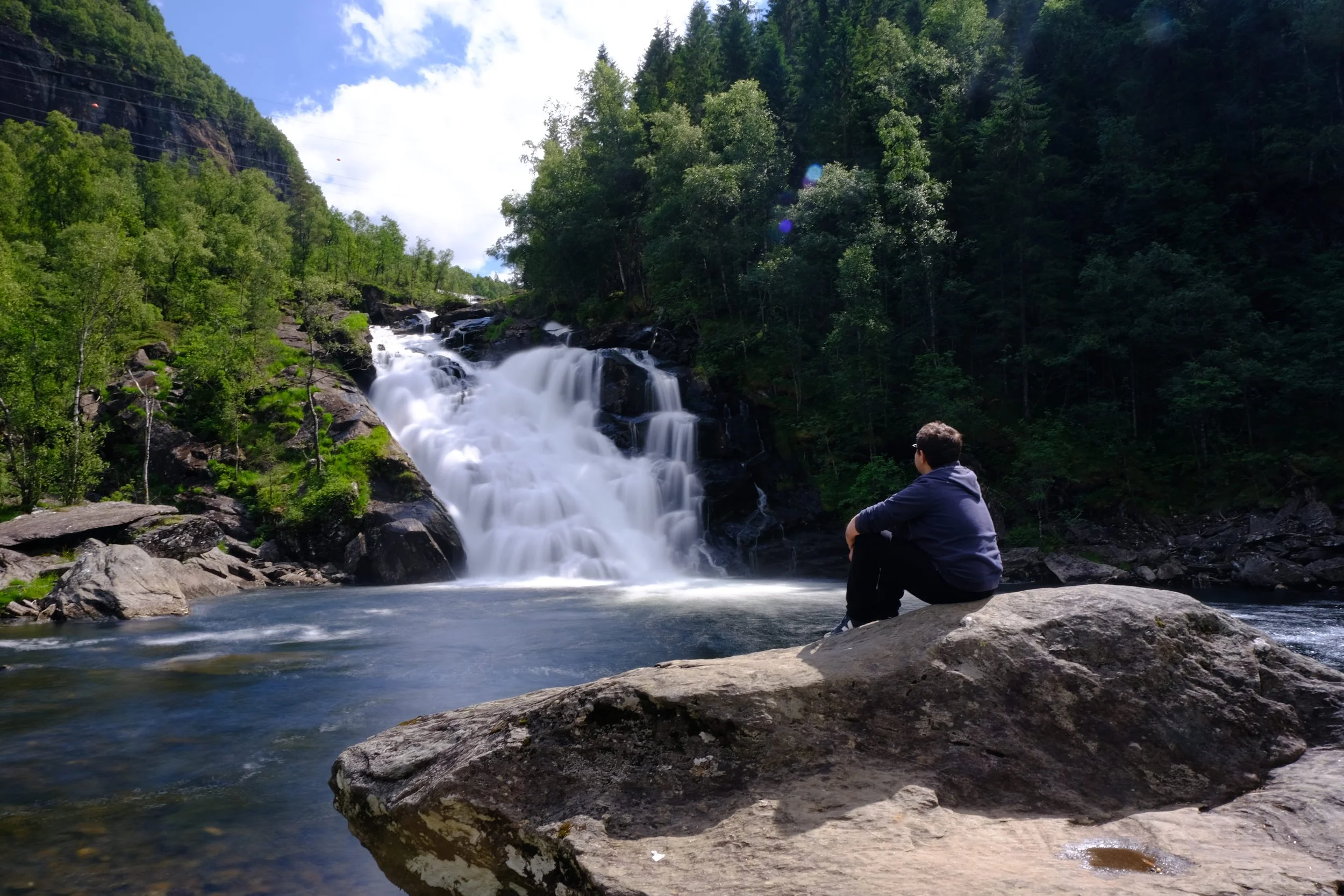Ein Mann sitzt auf einem großen Stein am Fluss und schaut auf einen Wasserfall inmitten eines grünen Waldes bei sonnigem Wetter.