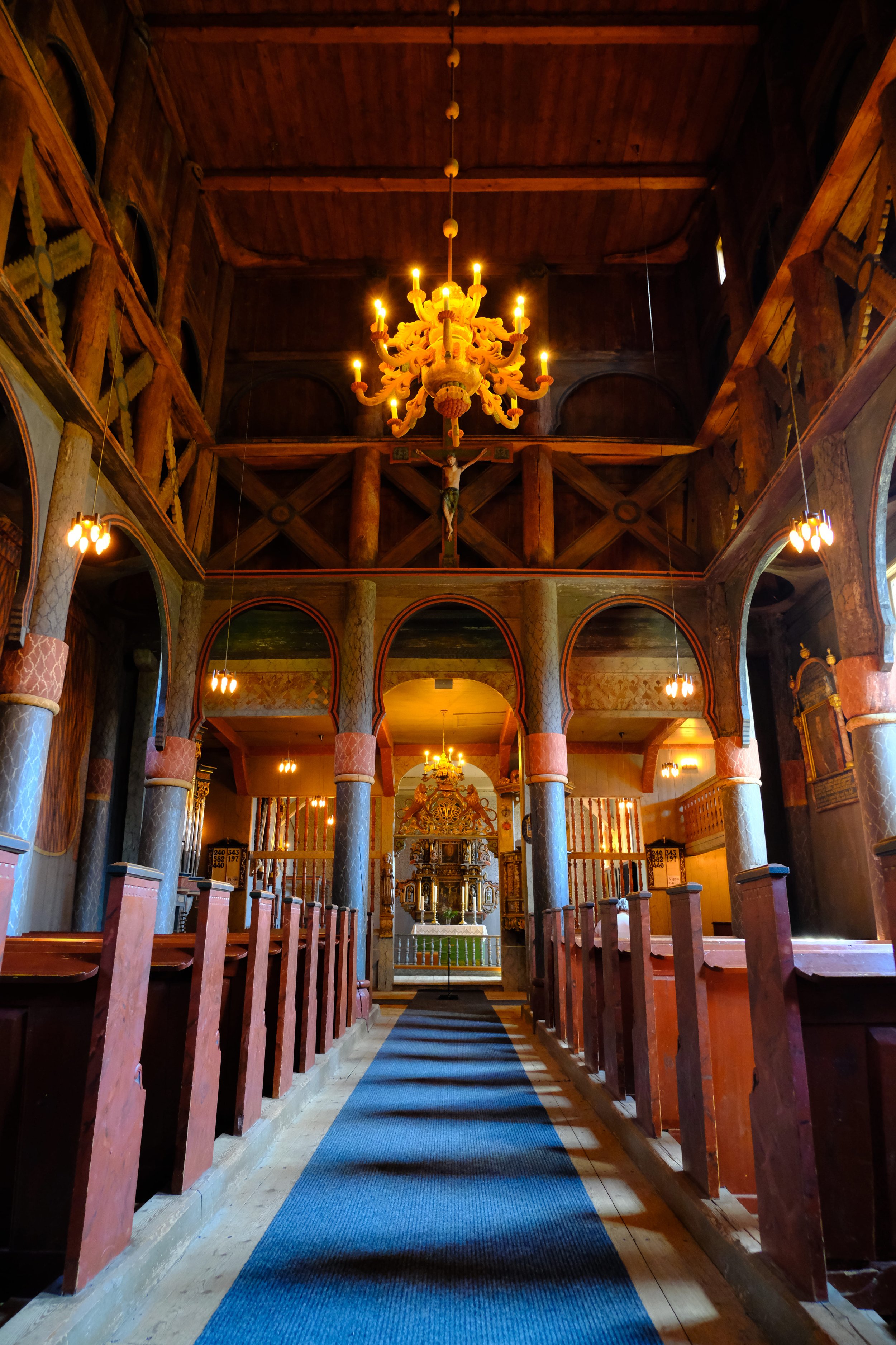 Innenraum einer Kirche mit Holzdekoration, einem Kronleuchter in der Mitte, Bänken auf beiden Seiten und einem Altar im Hintergrund.