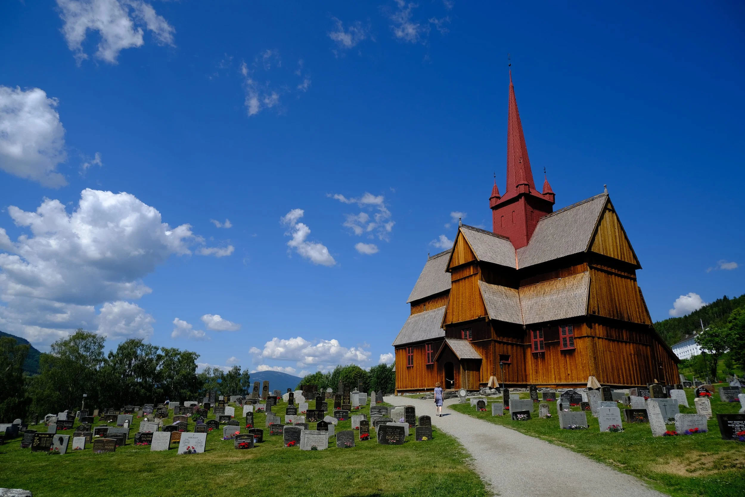 Ein großes Holzschiffkirche mit hohem roten Turm, umgeben von einem Friedhof mit Grabsteinen, bei schönem blauen Himmel.