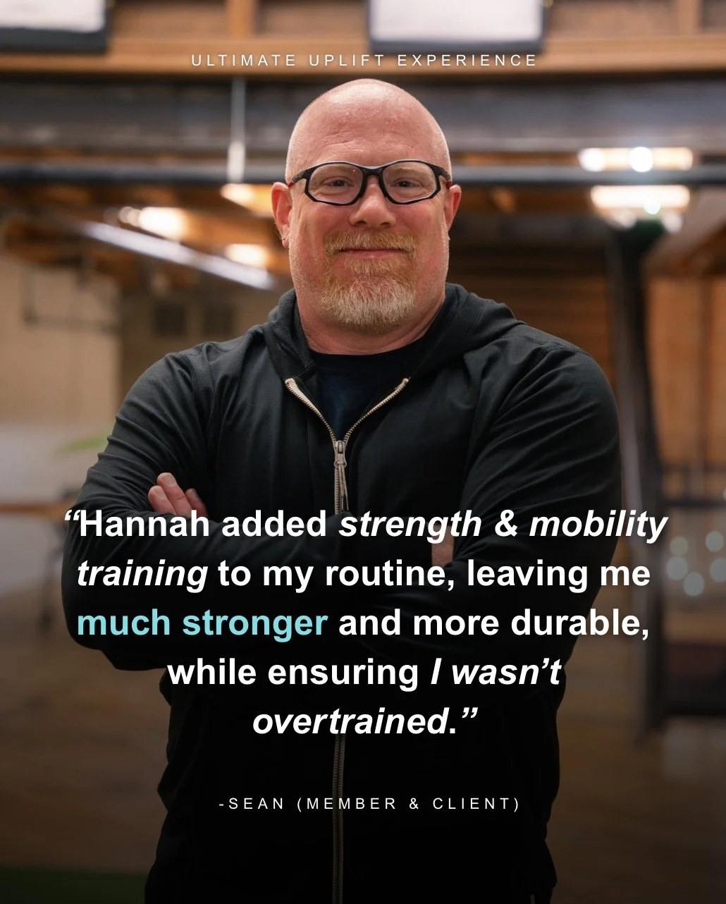 A man with glasses and a goatee smiling, standing with arms crossed in a gym or fitness space with wooden beams and bright lighting in the background.