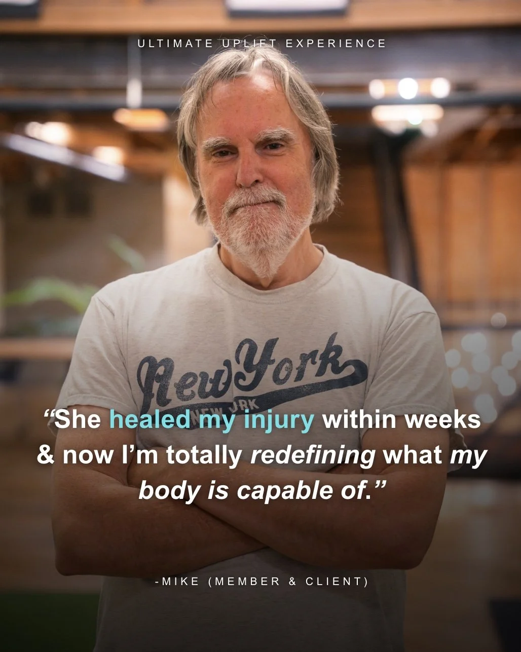 A smiling older man with gray hair and a beard, wearing a white T-shirt with 'New York' written on it, standing indoors with blurred background lights.