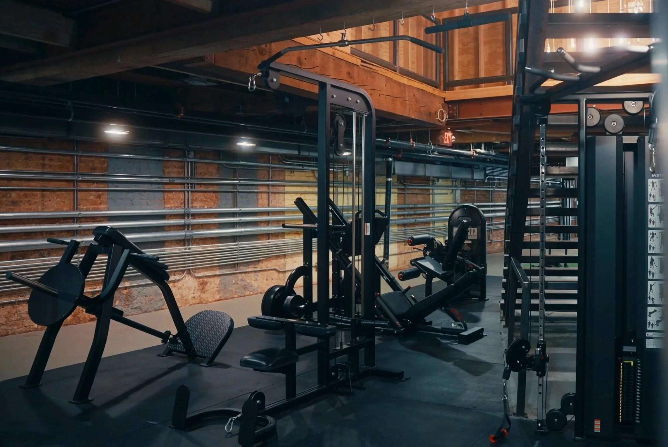 A woman with long, wavy blonde hair and a black workout outfit smiling with her hand on her hips in a gym with wooden wall panels, dumbbells, and a large mirror in the background.