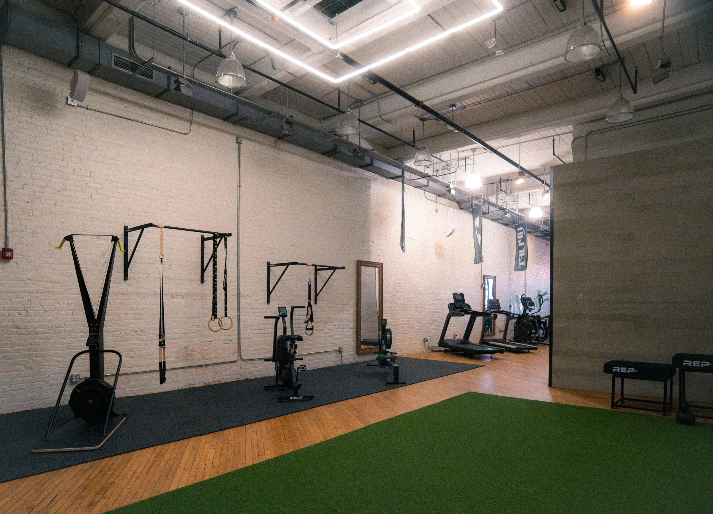 Interior of a gym with workout equipment including rowing machines, stationary bikes, and hanging gymnastic rings. The gym has a white brick wall, wooden flooring, and exposed ceiling with industrial lighting.