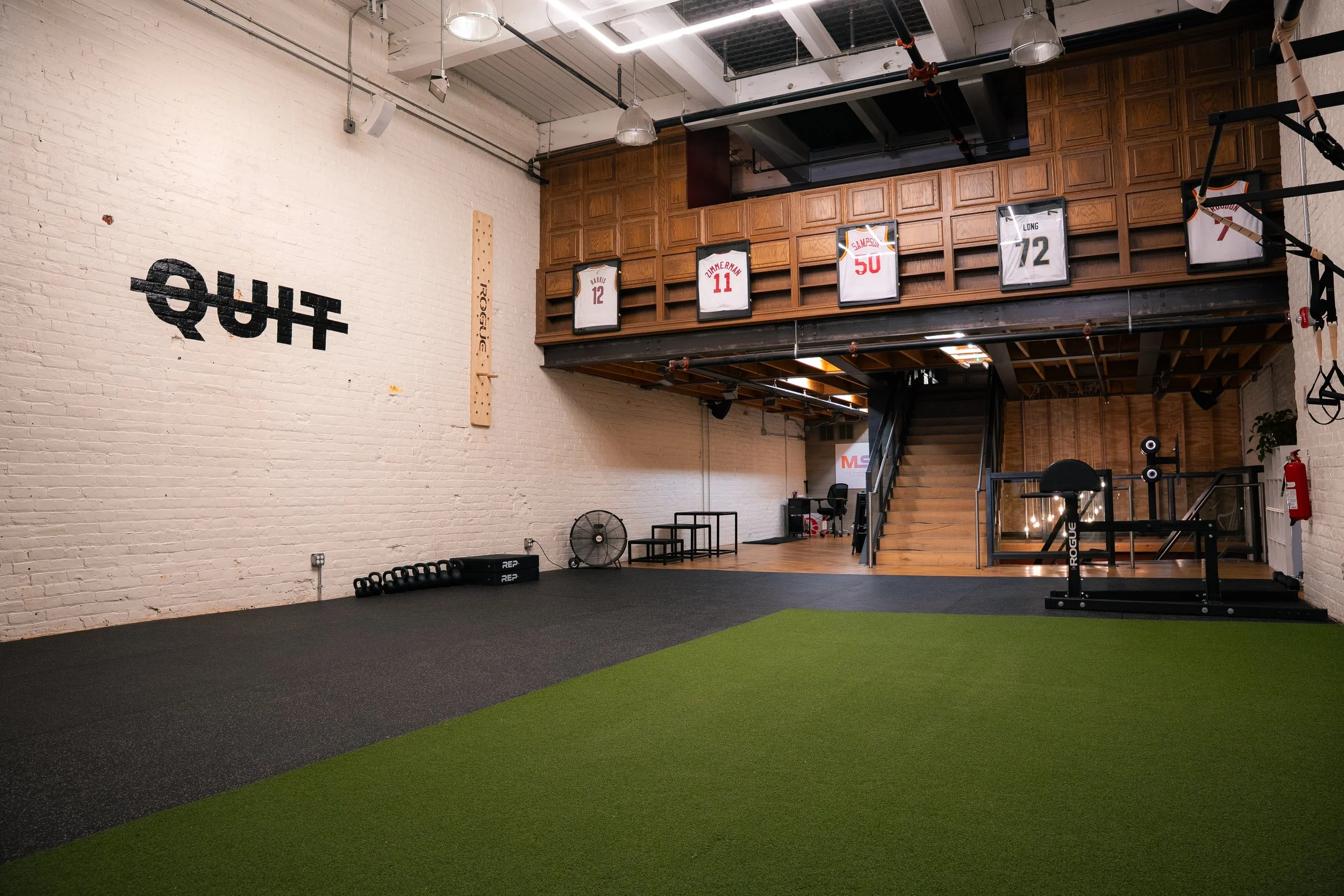 Woman riding a stationary exercise bike in a gym with a white brick wall and gym equipment hanging from the ceiling.