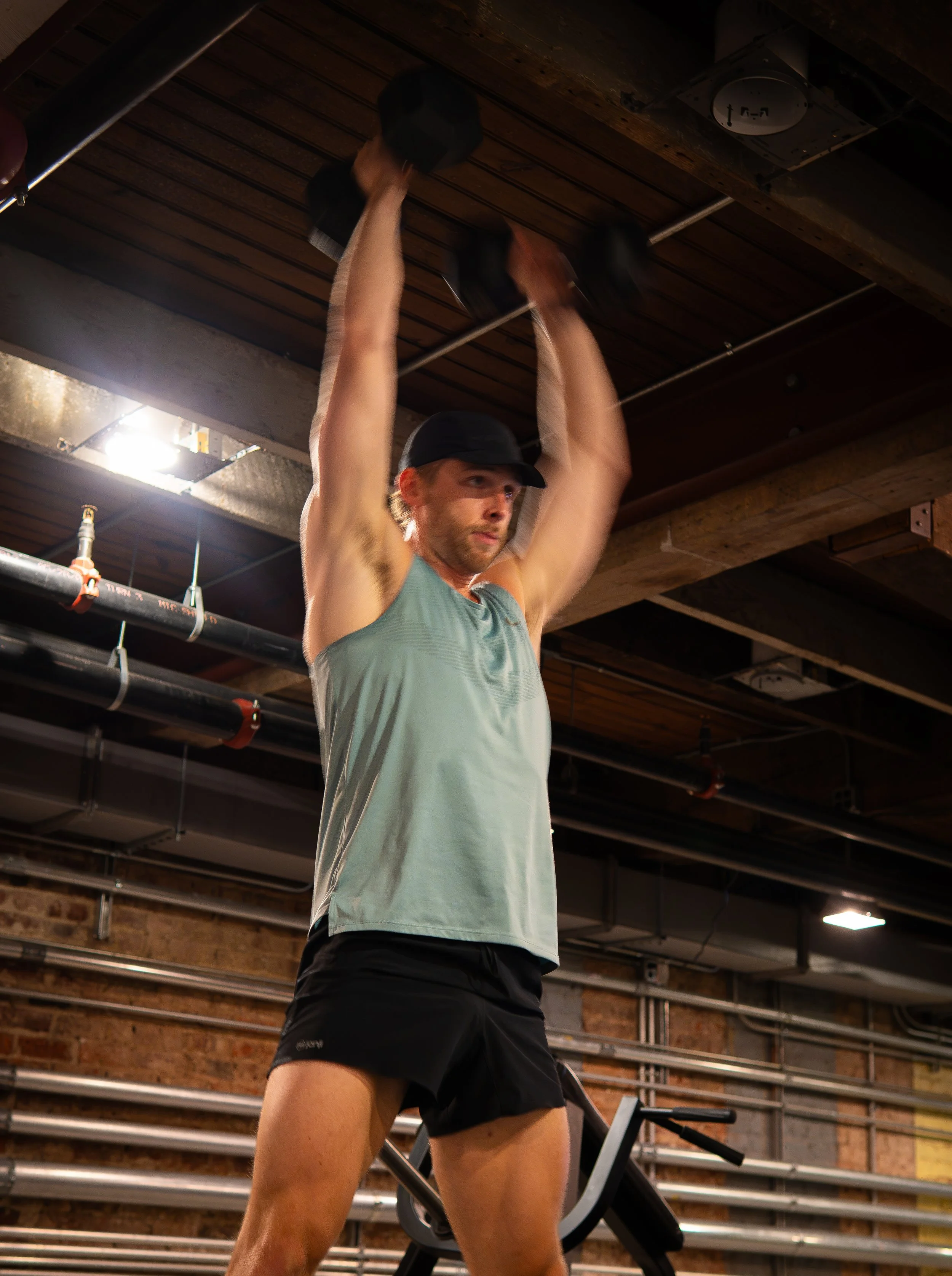 A man lifting a dumbbell overhead while standing in a gym with exposed brick walls and industrial ceiling pipes.