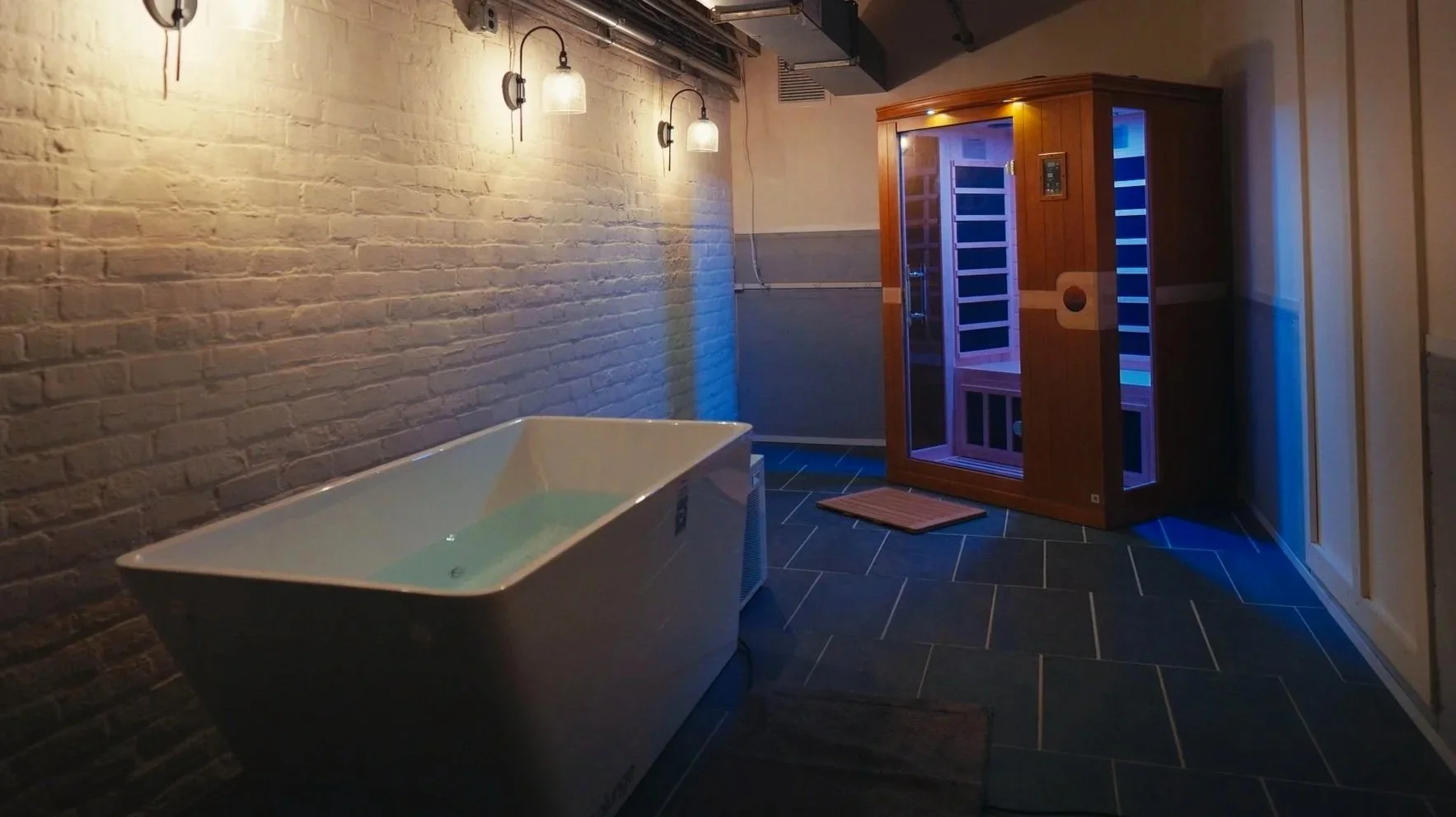 A woman with long blonde hair looking into a sauna room with wooden walls and black tiles. The room is illuminated with blue and purple lighting.