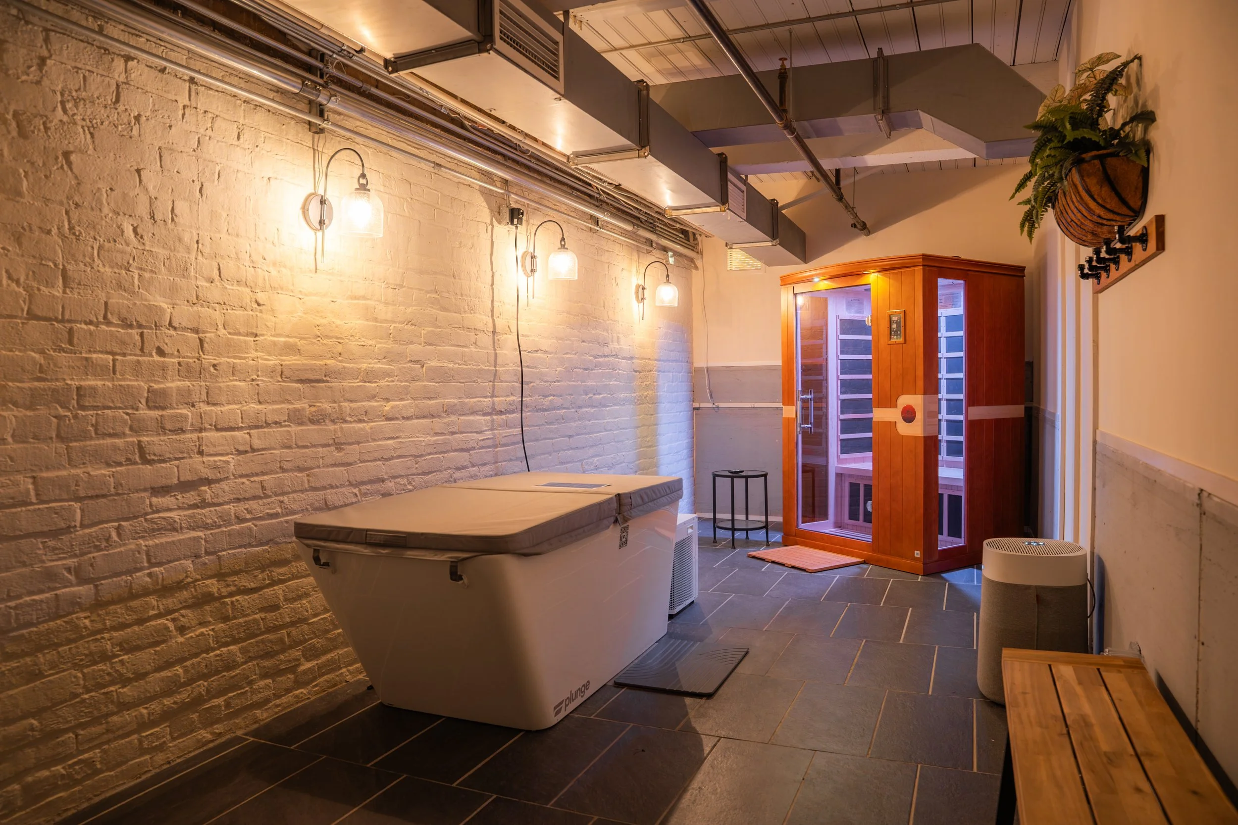 A room with a brick wall, a white bathtub, a wooden sauna, and a small seating area with a wooden bench and a cylindrical air purifier.