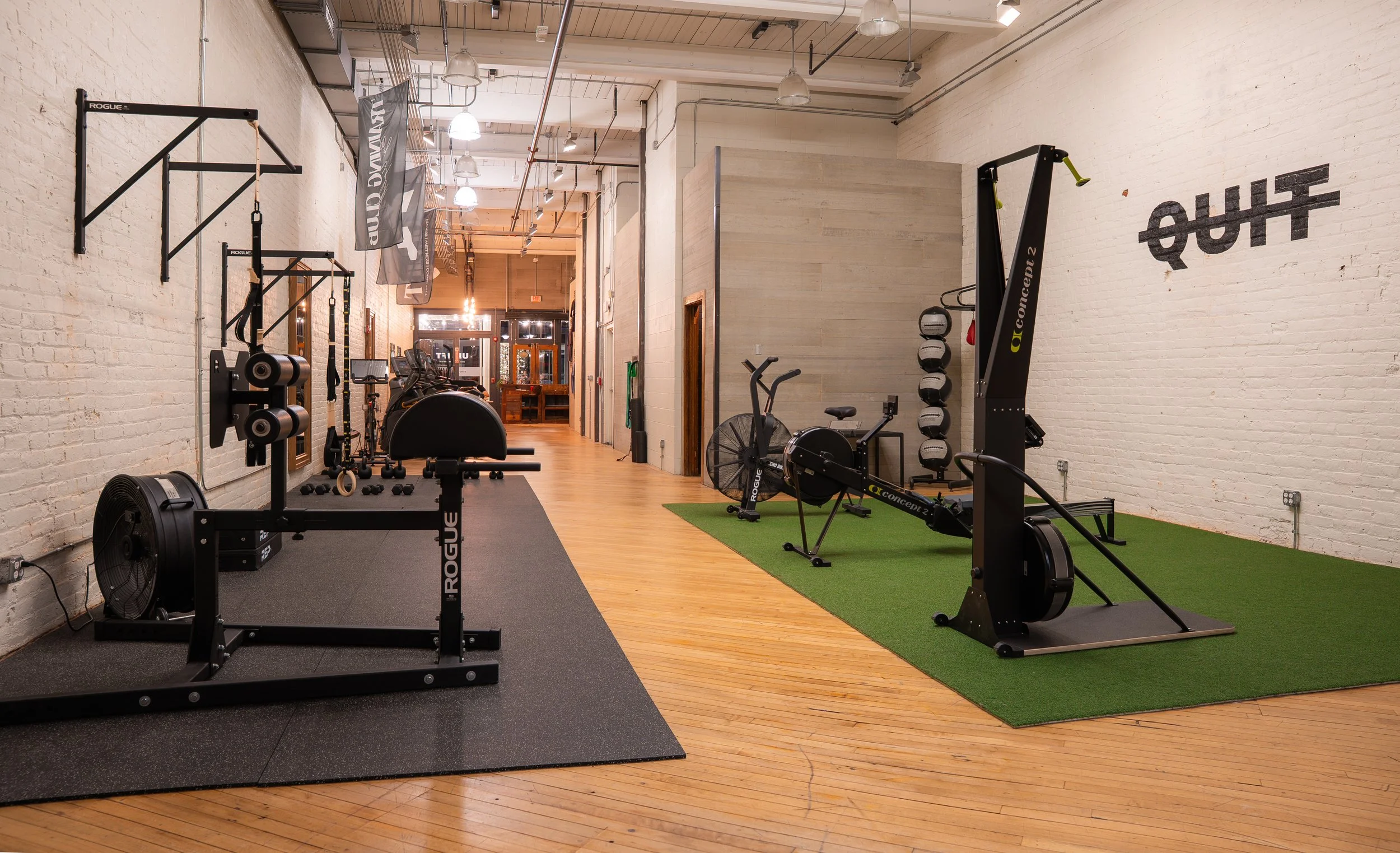 Woman riding a stationary exercise bike in a gym with a white brick wall and gym equipment hanging from the ceiling.