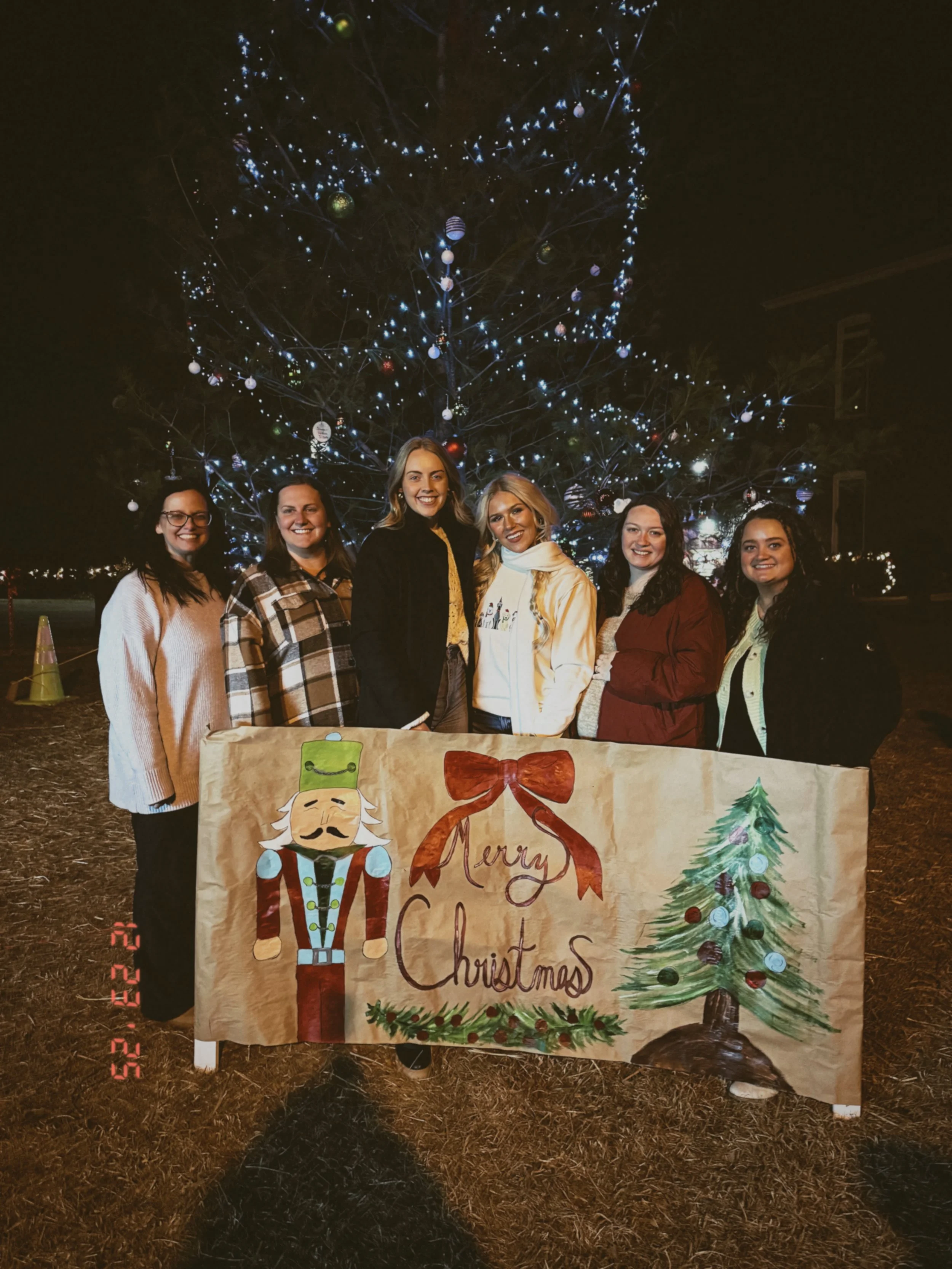 Group of six women standing outdoors at night in front of a decorated Christmas tree, holding a large banner with holiday illustrations and the words 'Merry Christmas'.