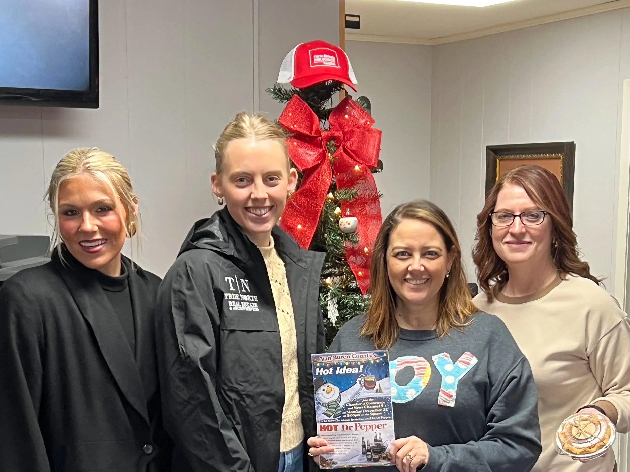 Four women standing in front of a decorated Christmas tree with a red bow and a red cap on top, smiling at the camera. One woman holds a magazine titled 'Hot Dr. Pepper'.
