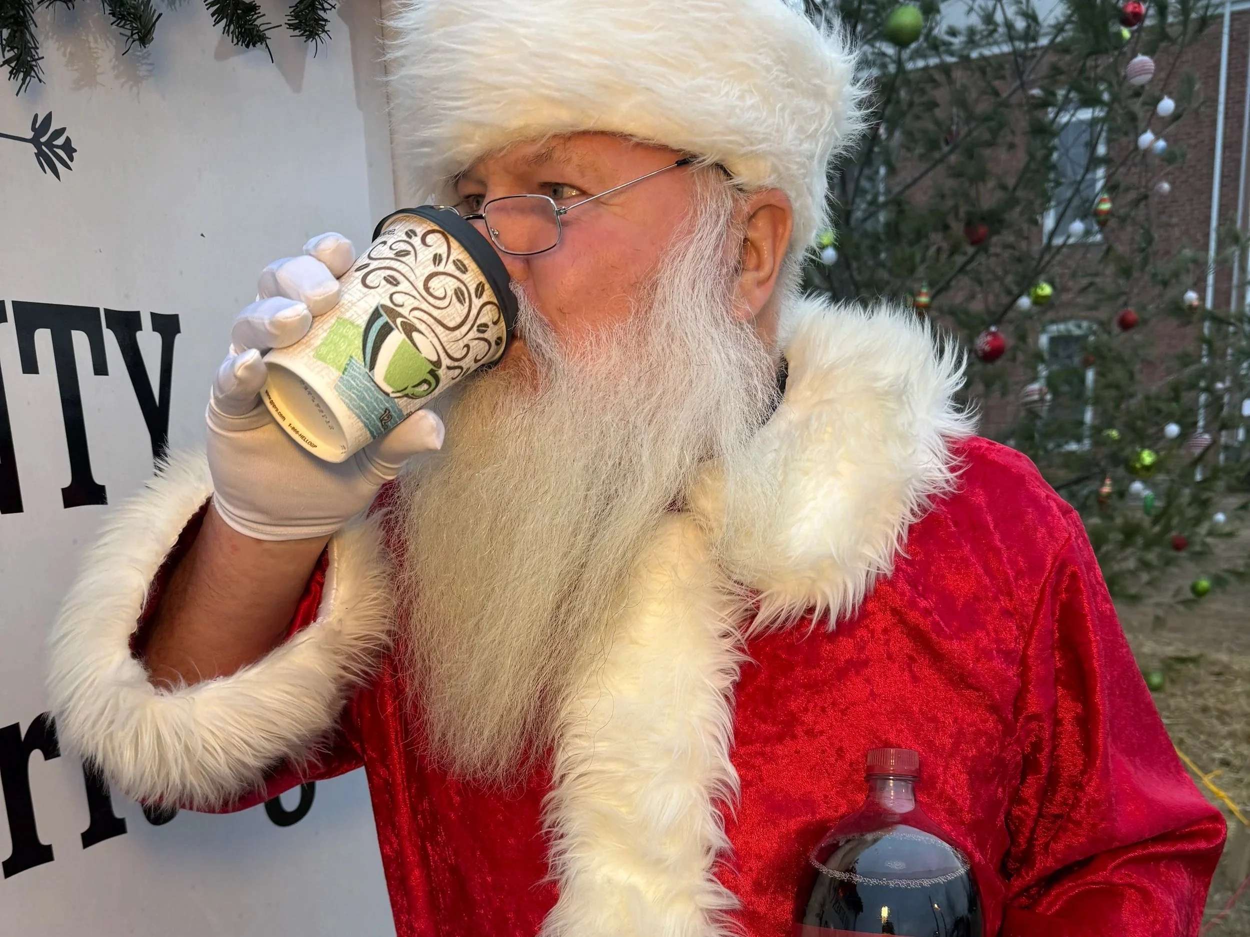 Santa Claus drinking from a paper cup outdoors, holding a bottle of soda, with a Christmas tree decorated with ornaments in the background.