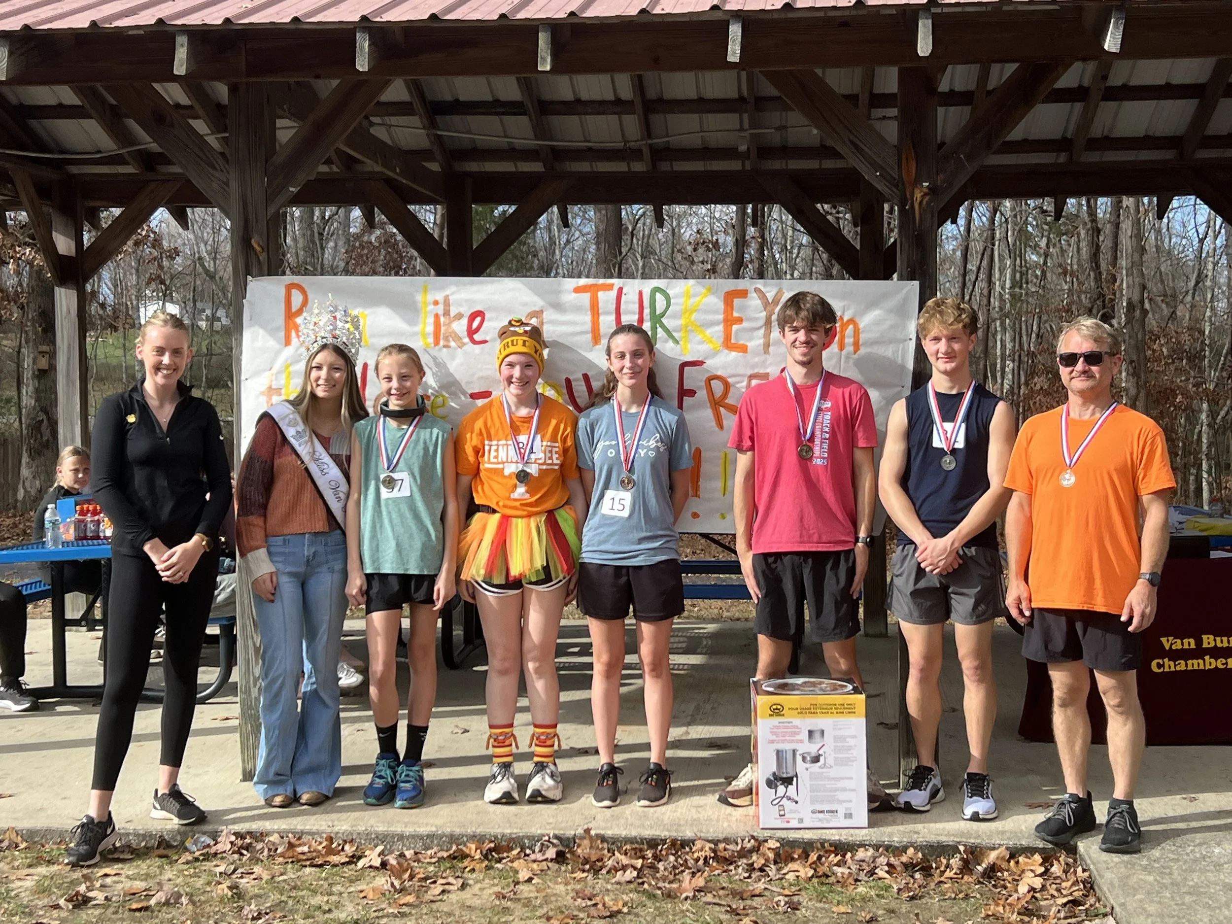 Group of people standing outdoors under a wooden pavilion, some wearing medals, celebrating a race event, with a colorful banner in the background.