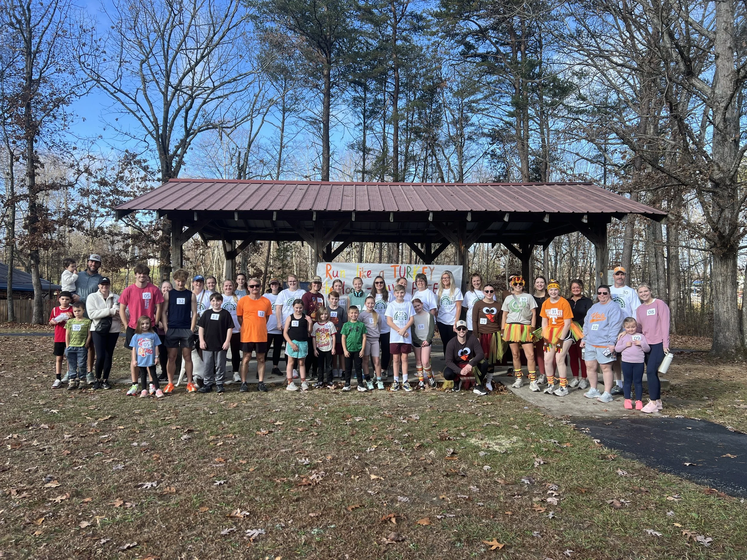 Group of people of various ages gathered outdoors in front of a pavilion for a running event, with a banner reading "Run like a Turkey" behind them. Some individuals are dressed in colorful and festive outfits, and there are children and adults in th
