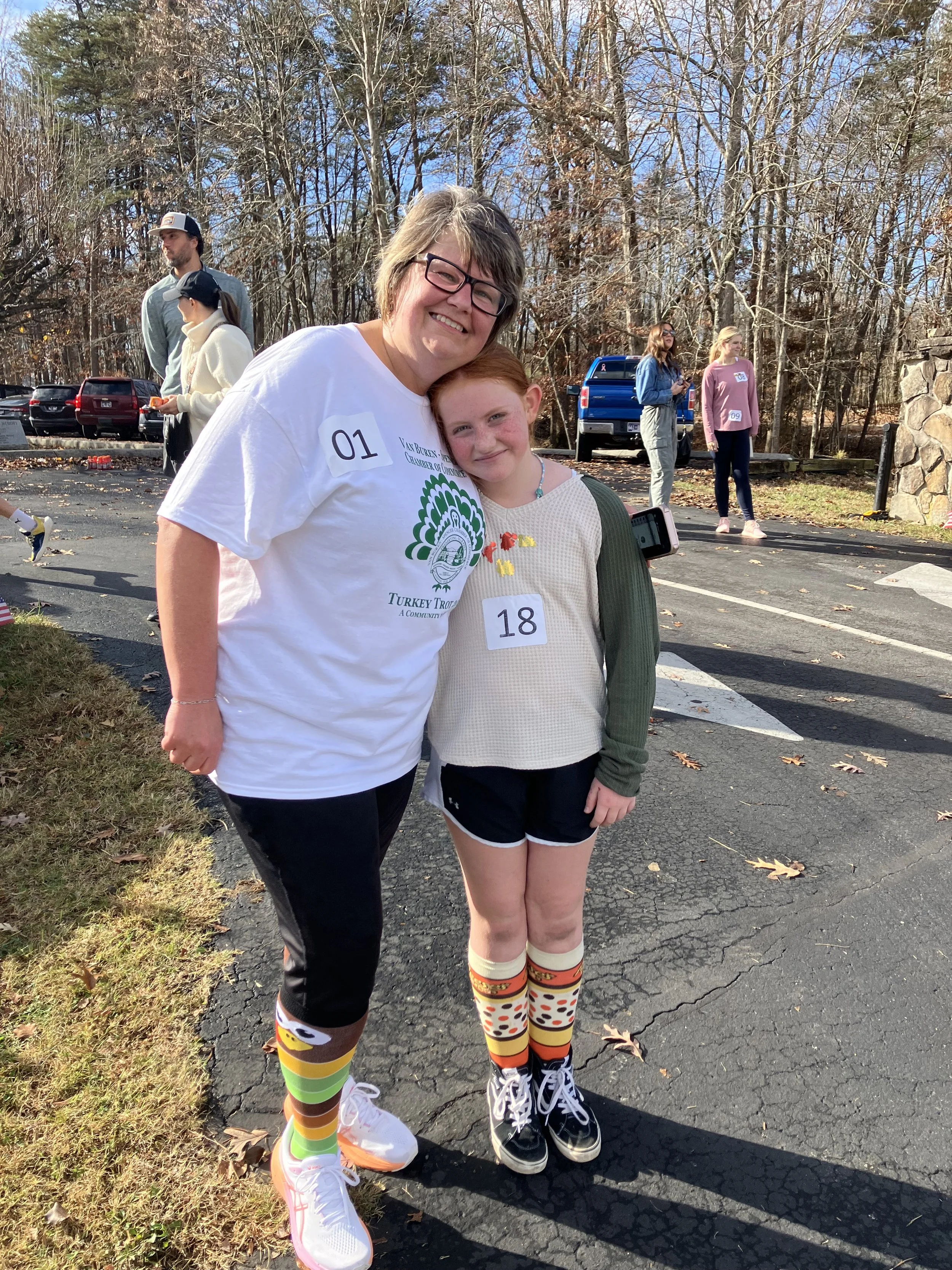 A woman and a young girl smile and pose together outdoors at an event, with other people in the background. The woman is wearing a white T-shirt, black pants, and colorful knee-high socks featuring a rainbow and smiley face design. The girl is wearin