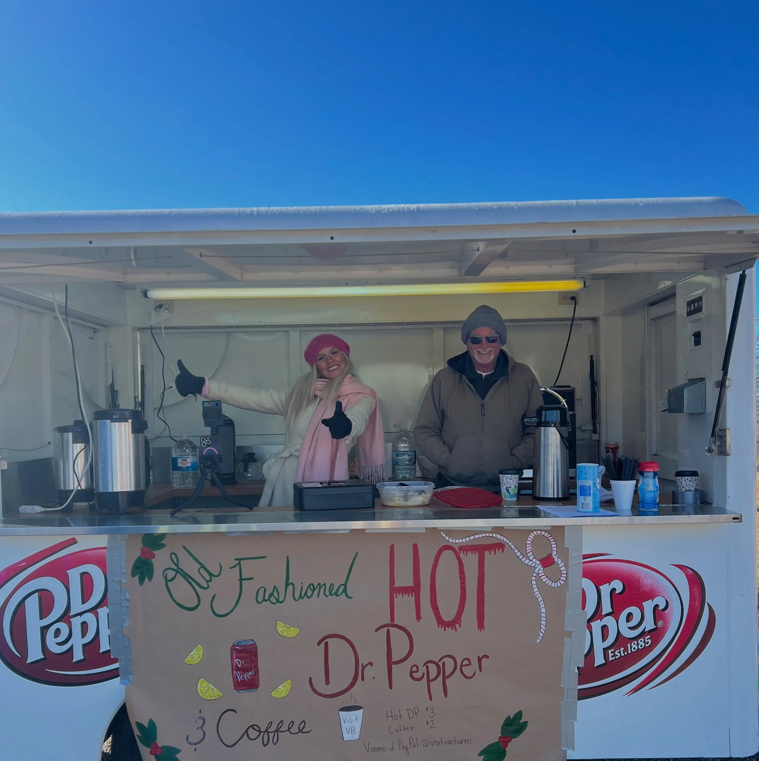A woman and man standing in a mobile coffee stand, smiling and posing, with a sign reading "Old Fashioned HOT Dr. Pepper." The woman is wearing a pink hat, white coat, and scarf, and the man is wearing a gray beanie and sunglasses. The stand has vari