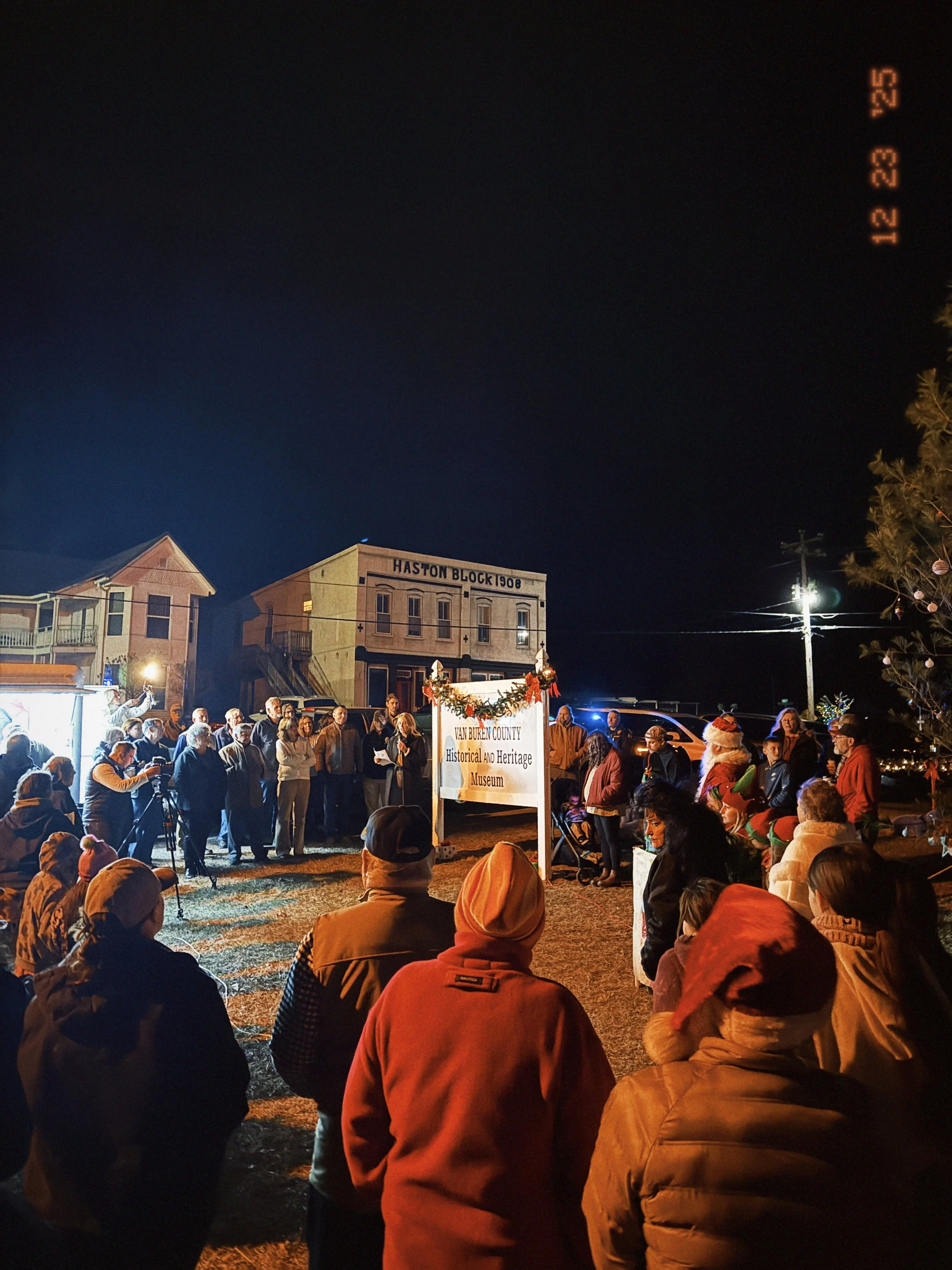 Community gathering at night in front of the Van Buren County Historical and Heritage Museum, decorated for Christmas, with people including Santa Claus, a Christmas tree, and a group of spectators.