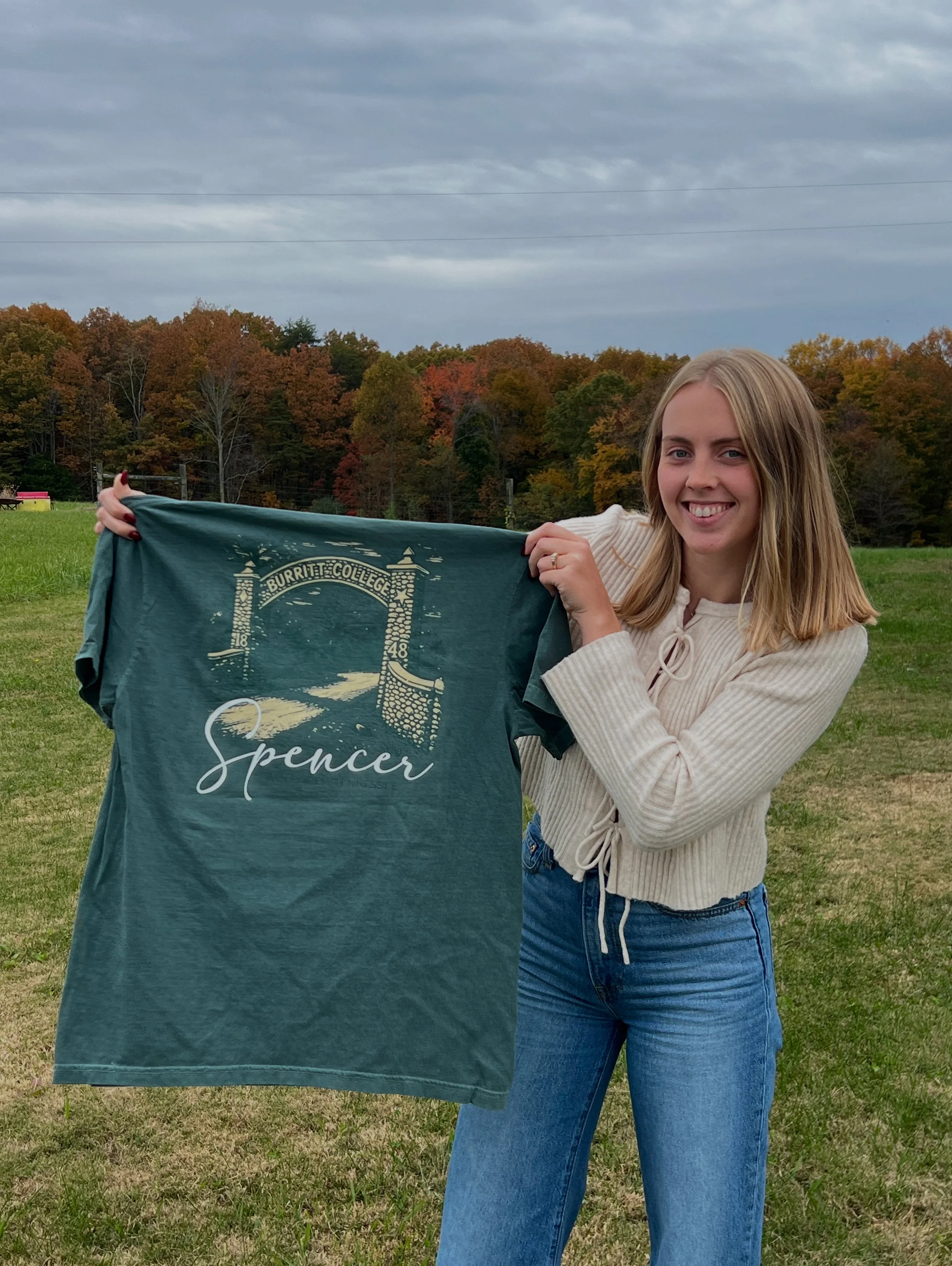 A young woman holding a faded green T-shirt with a design of Burritt College archway and the word "Spencer" on it, standing in an open grassy field with trees showing fall colors in the background.