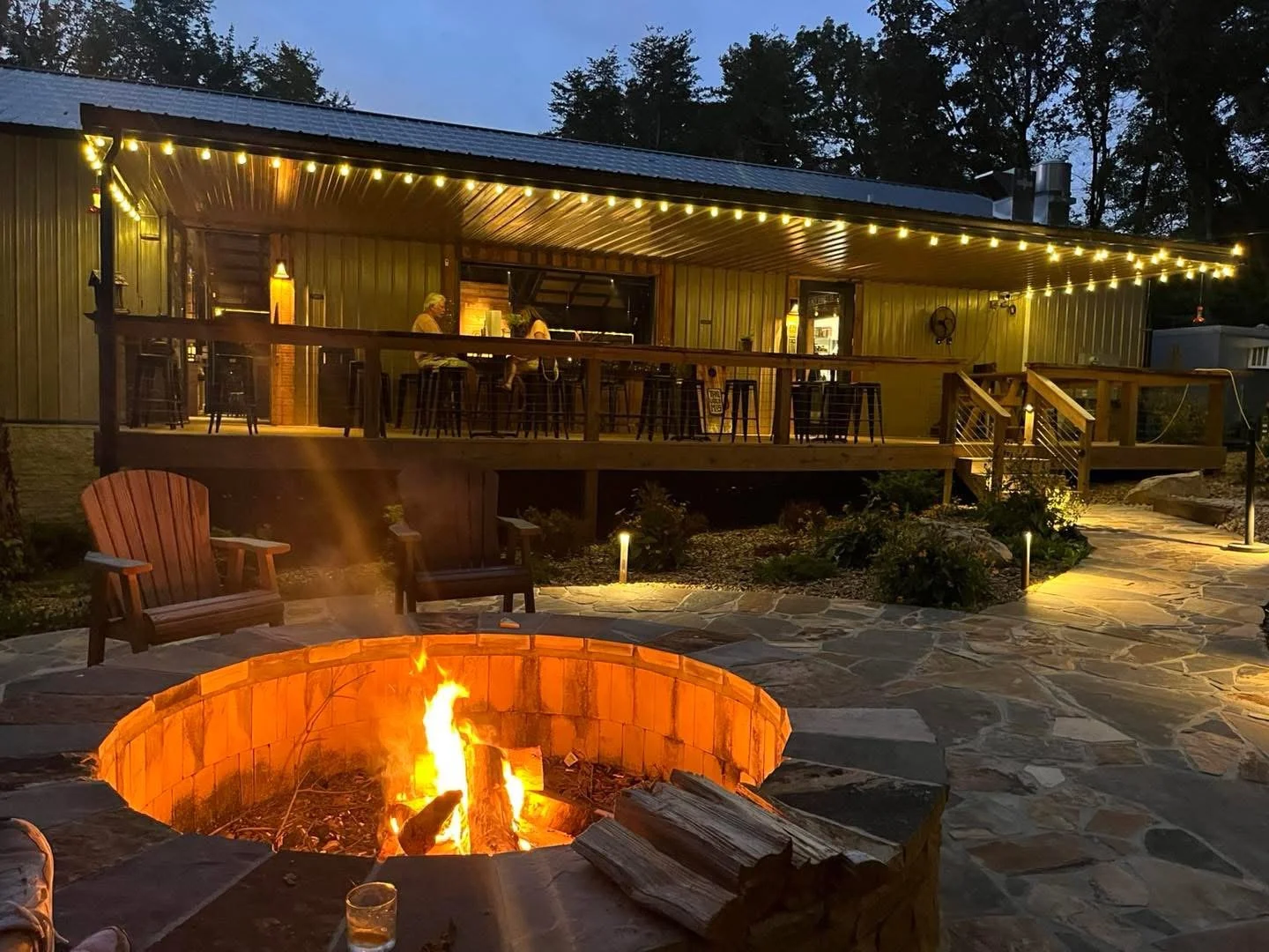 An outdoor patio area with a fire pit in the foreground, wood chairs, and a building with string lights overhead, with some people sitting inside at a bar or counter.