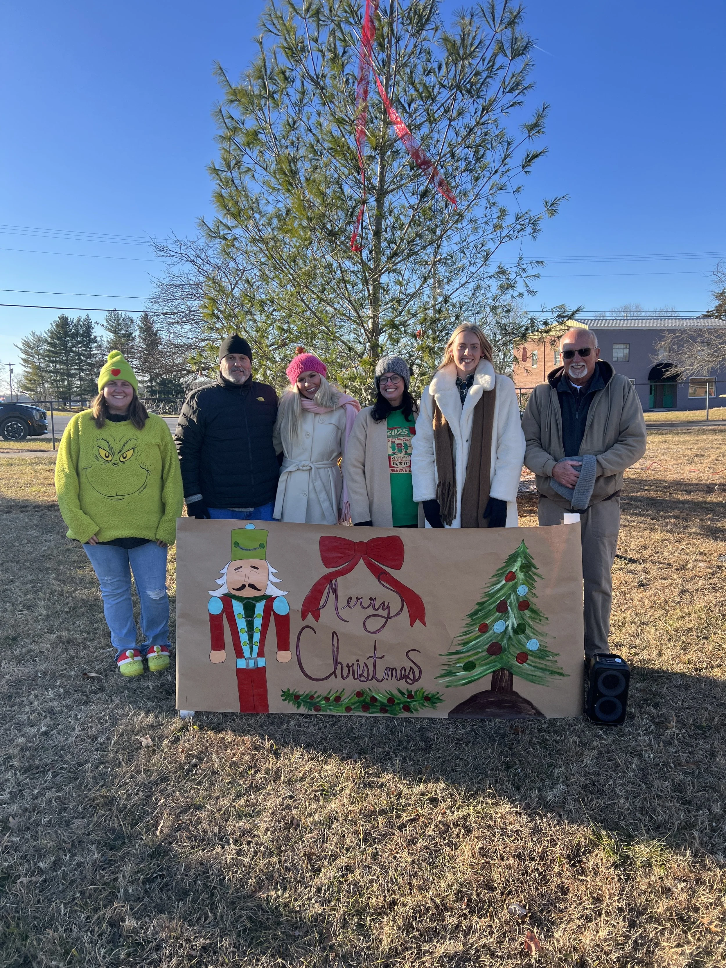 Group of six people standing outdoors during winter, holding a 'Merry Christmas' banner in front of a decorated Christmas tree, with a clear blue sky and residential houses in the background.