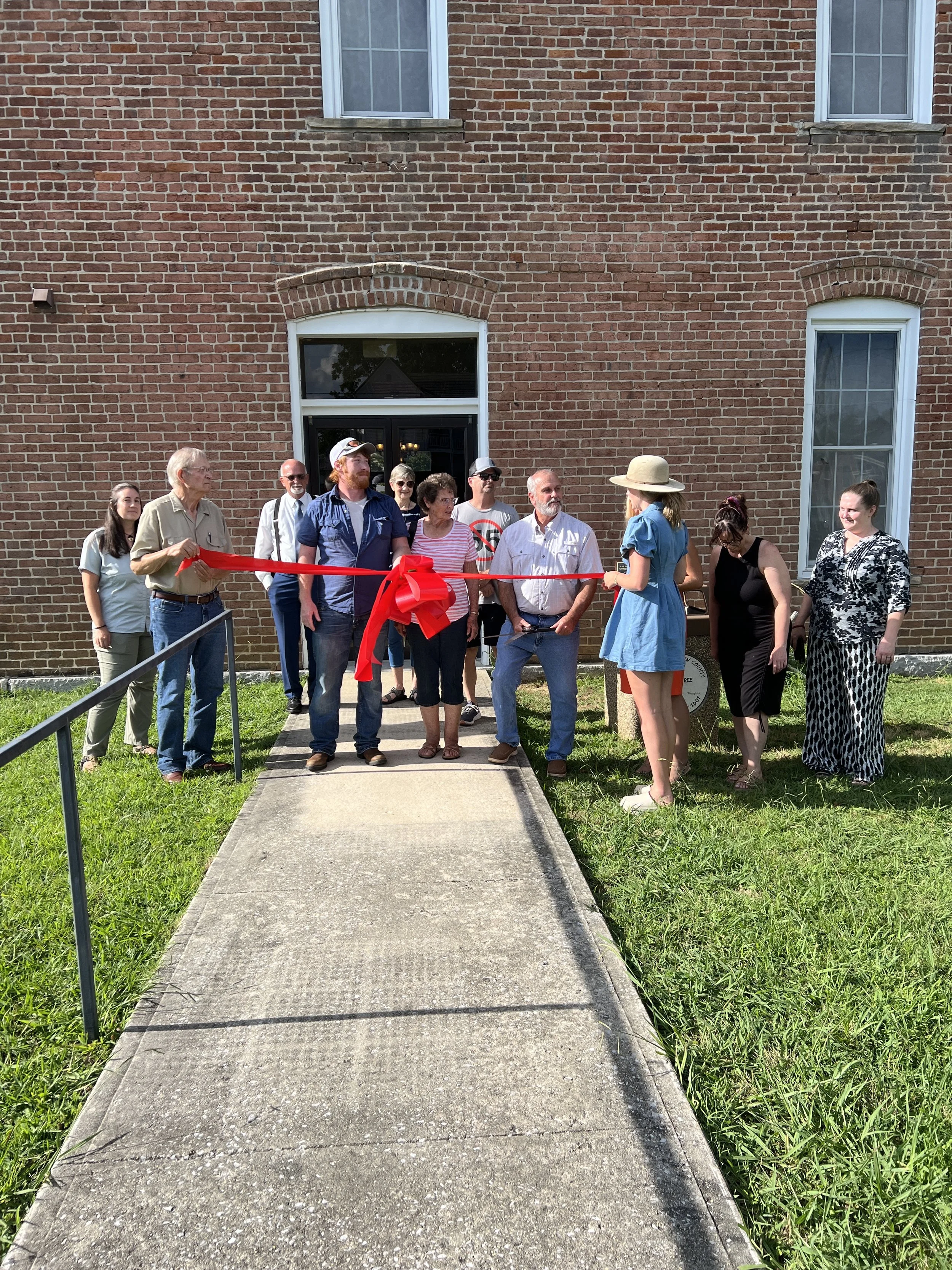 A ribbon-cutting ceremony with a group of people gathered outside a brick building. A woman with a large sunhat is holding scissors, preparing to cut a red ribbon while others watch.