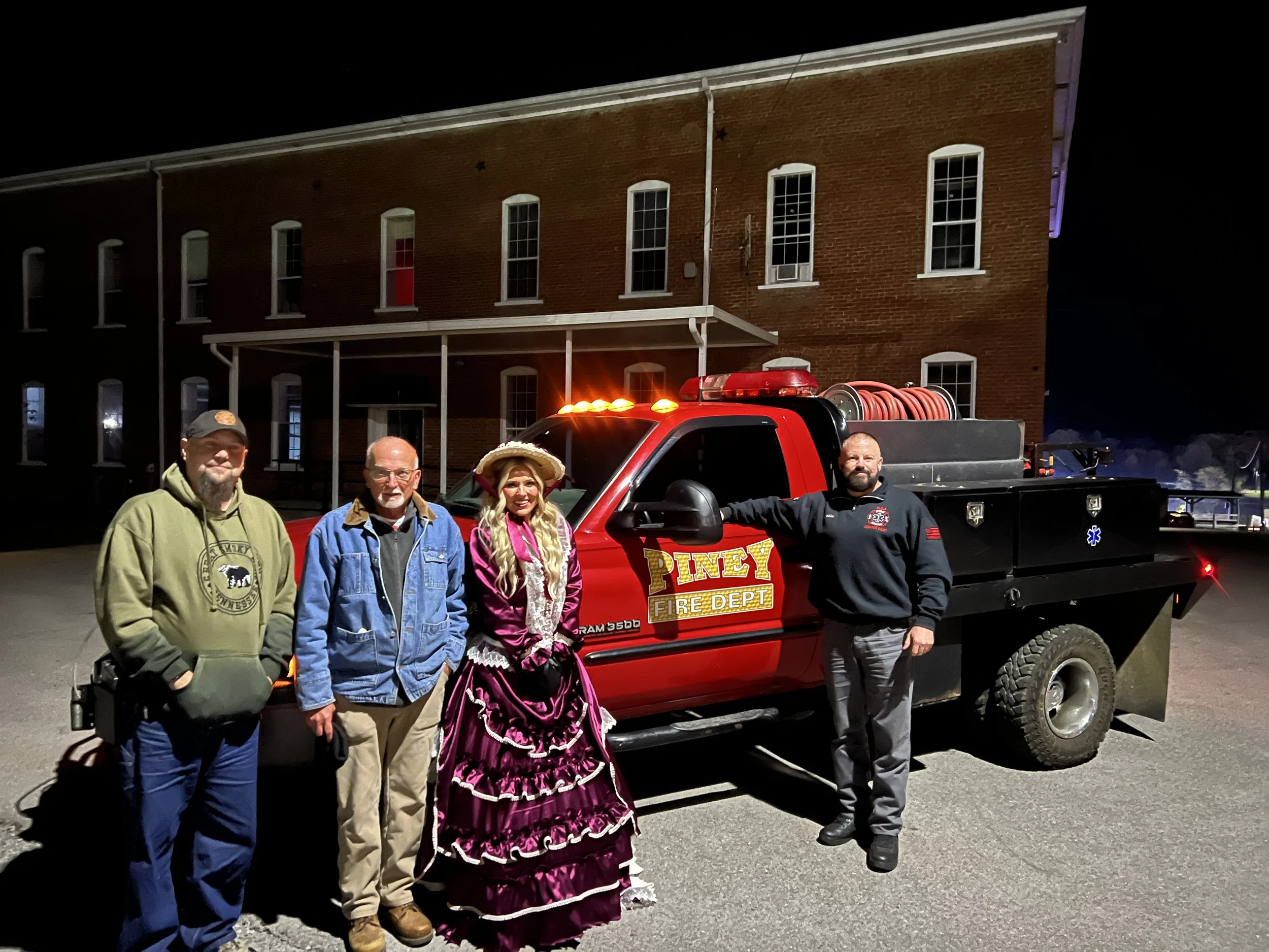 Four people stand in front of a red fire department truck with the sign 'Piney Fire Dept.' at night, with a brick building in the background. Two men are dressed casually, one woman wears vintage dress and hat, and the other man is in a firefighter u