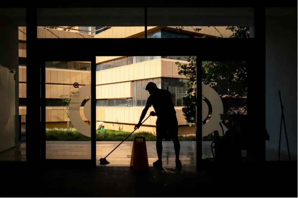A silhouette of a person mopping the floor inside a building with glass doors, with a modern building visible outside.
