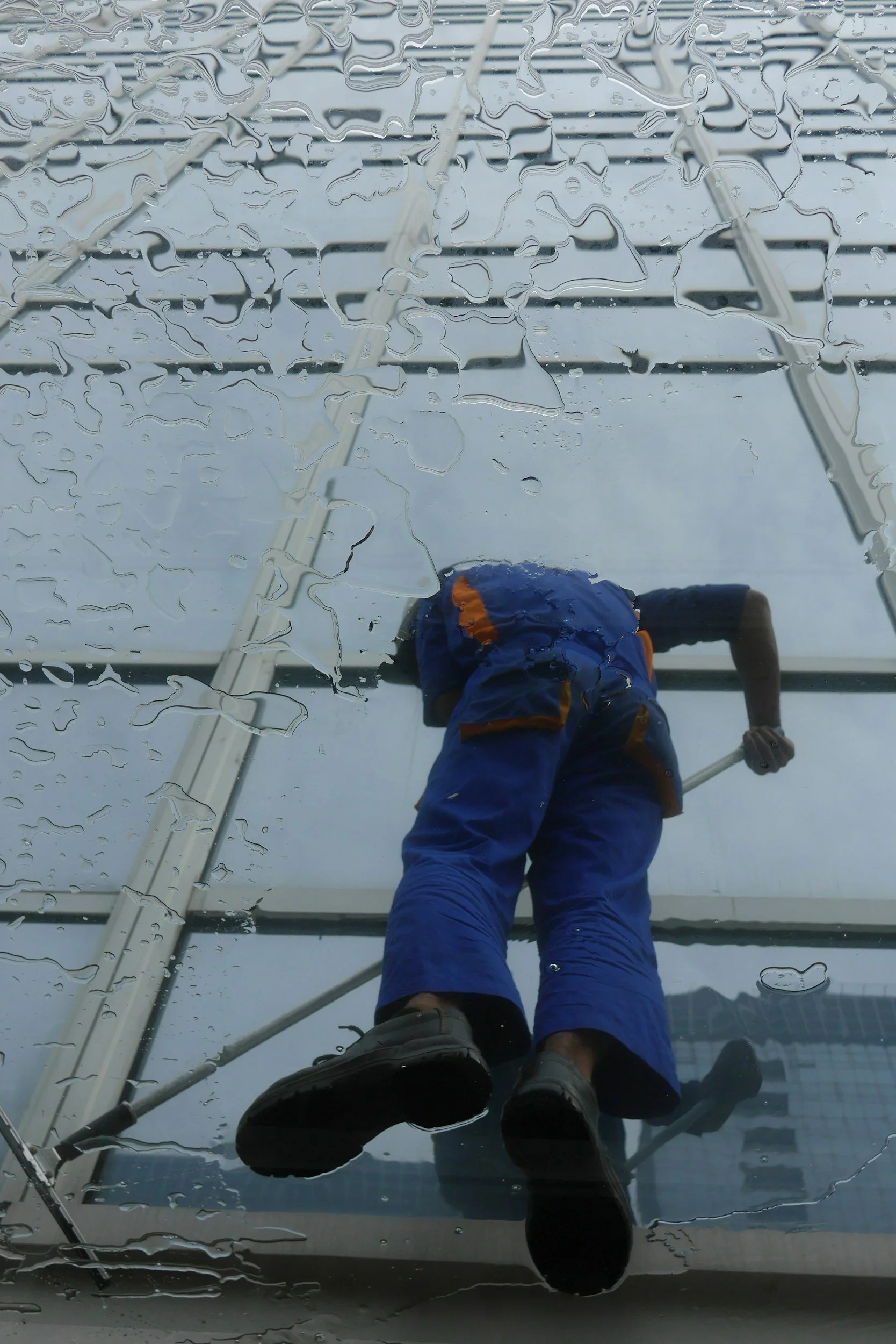 A person in blue outdoor gear and gray shoes is cleaning or inspecting a glass surface, with a reflection showing a ladder, a person, and a cloudy sky with raindrops.