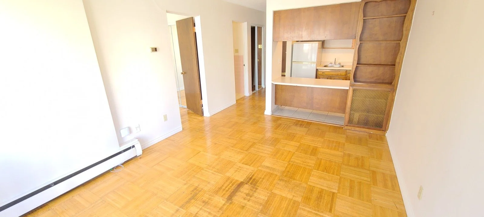 Empty living room with hardwood floor, white walls, open doorway leading to kitchen with wooden cabinets and a refrigerator. Built-in wooden shelves and cabinet are on right side.