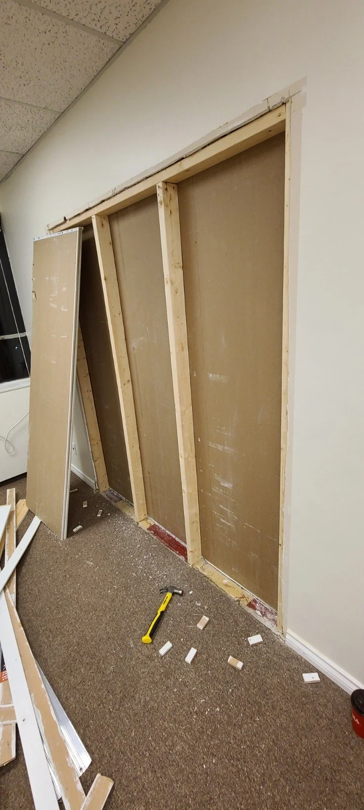Construction work in progress showing a partially installed wall frame with three vertical studs and drywall panels leaning against the wall, along with a hammer on the floor surrounded by small construction debris.