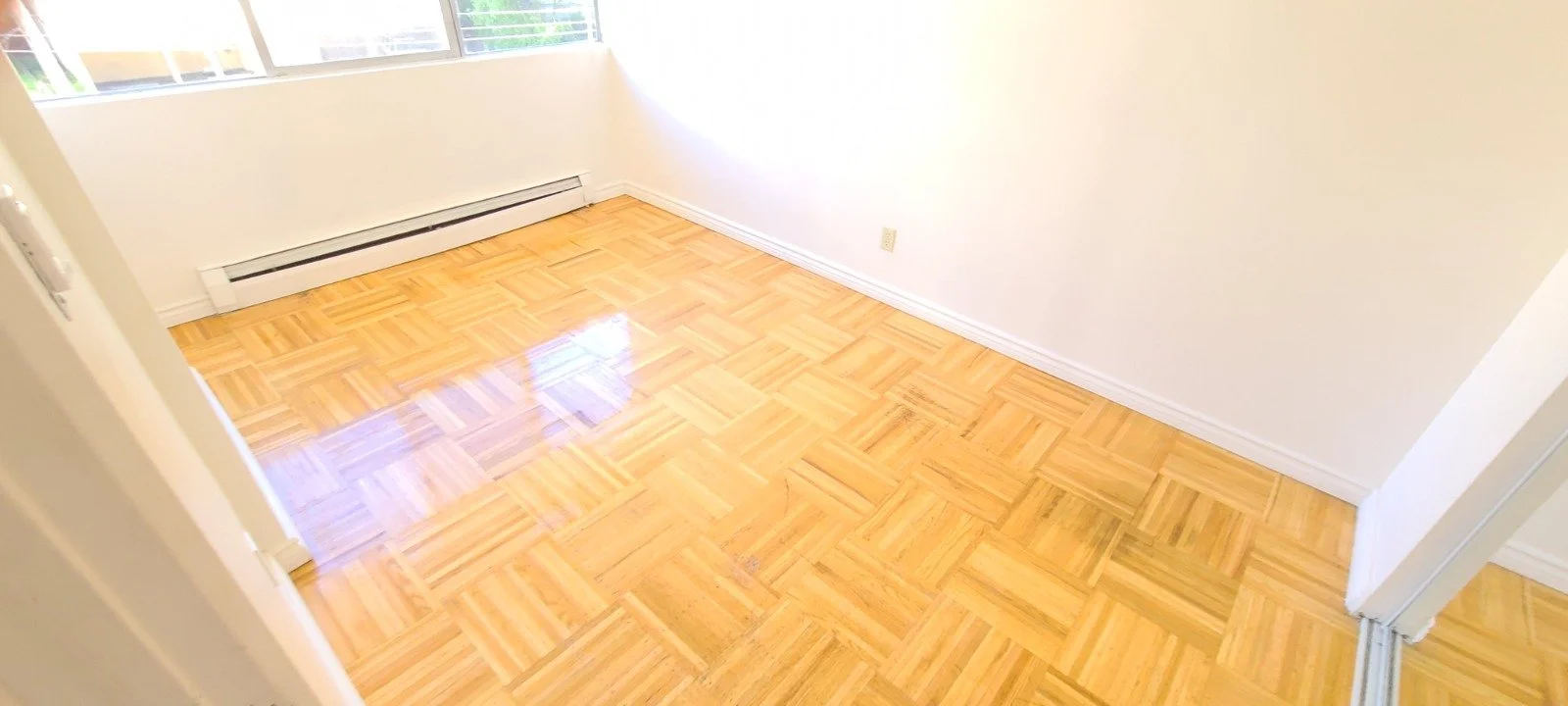Empty room with light-colored walls, hardwood parquet flooring, large window, and baseboard heater.
